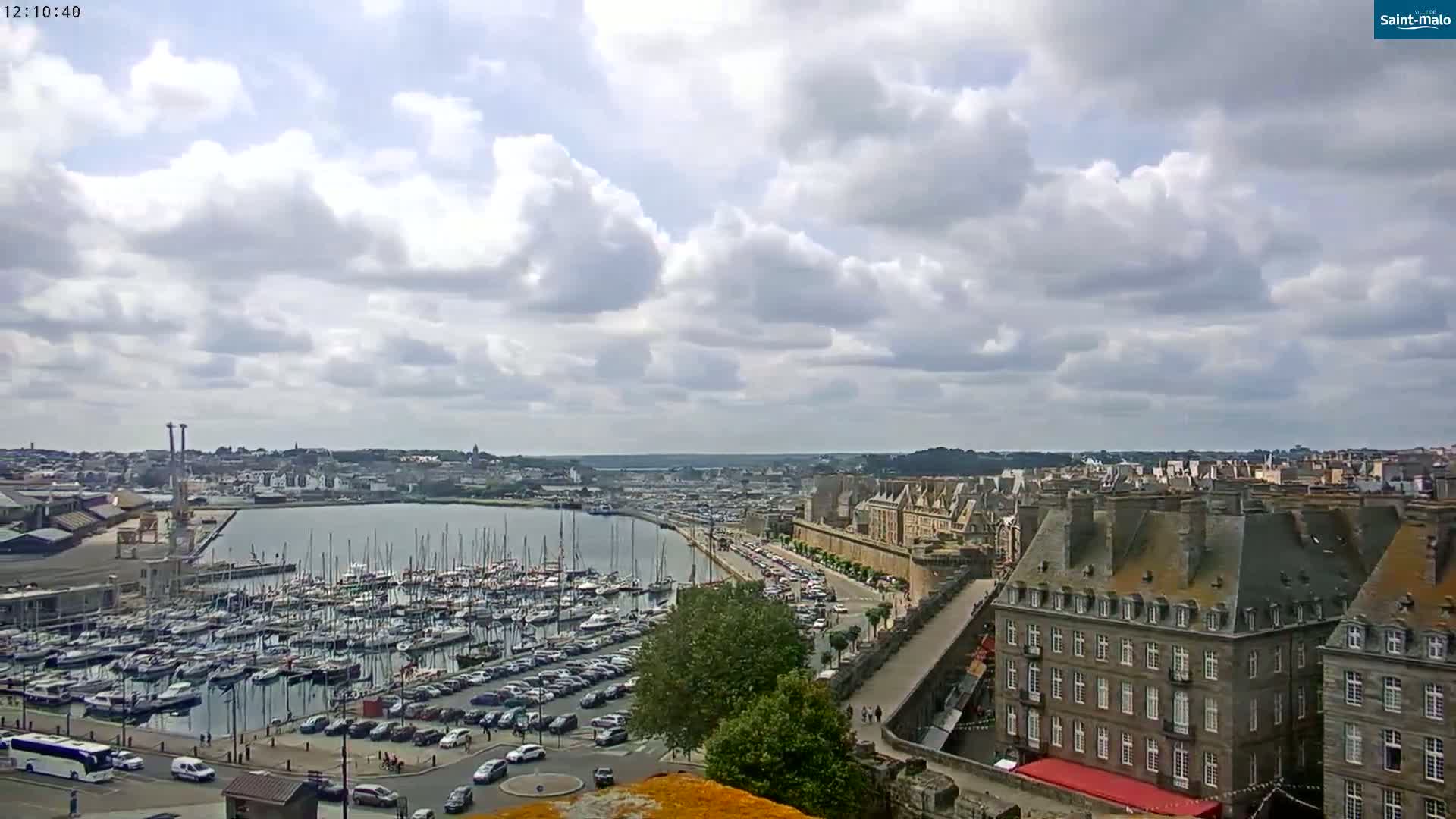 A partly cloudy day overlooks a harbor filled with sailboats, a city with stone buildings, and a parking lot.
