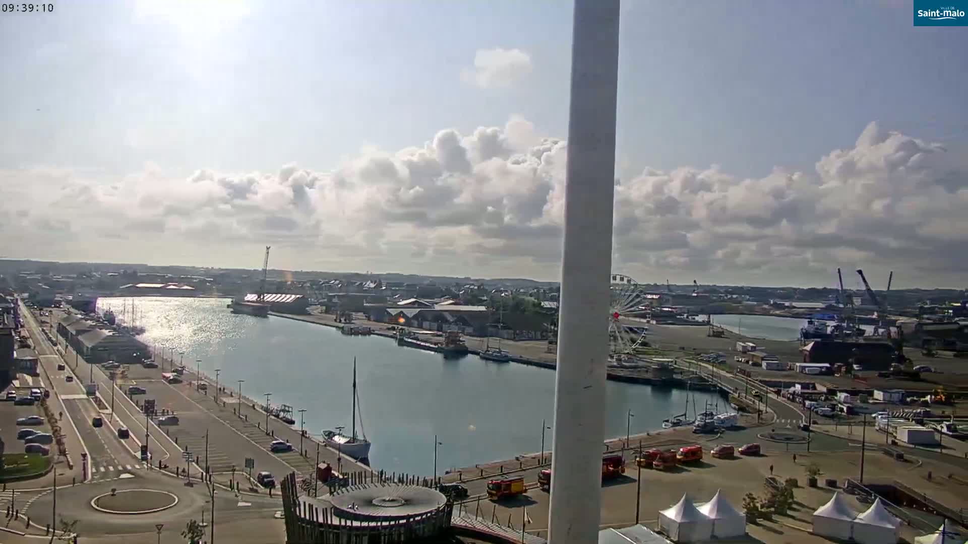 A partly sunny day shows a harbor with a Ferris wheel, boats, and buildings; a roadway is visible along the harbor's edge.