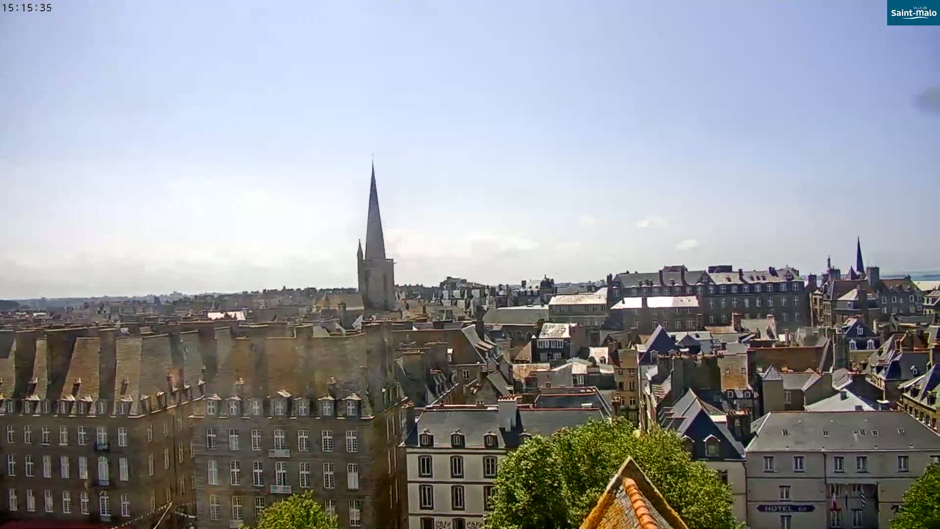A sunny day reveals a panoramic view of a European city, with numerous buildings of varying heights and styles, a prominent church spire in the background, and some green foliage in the foreground.