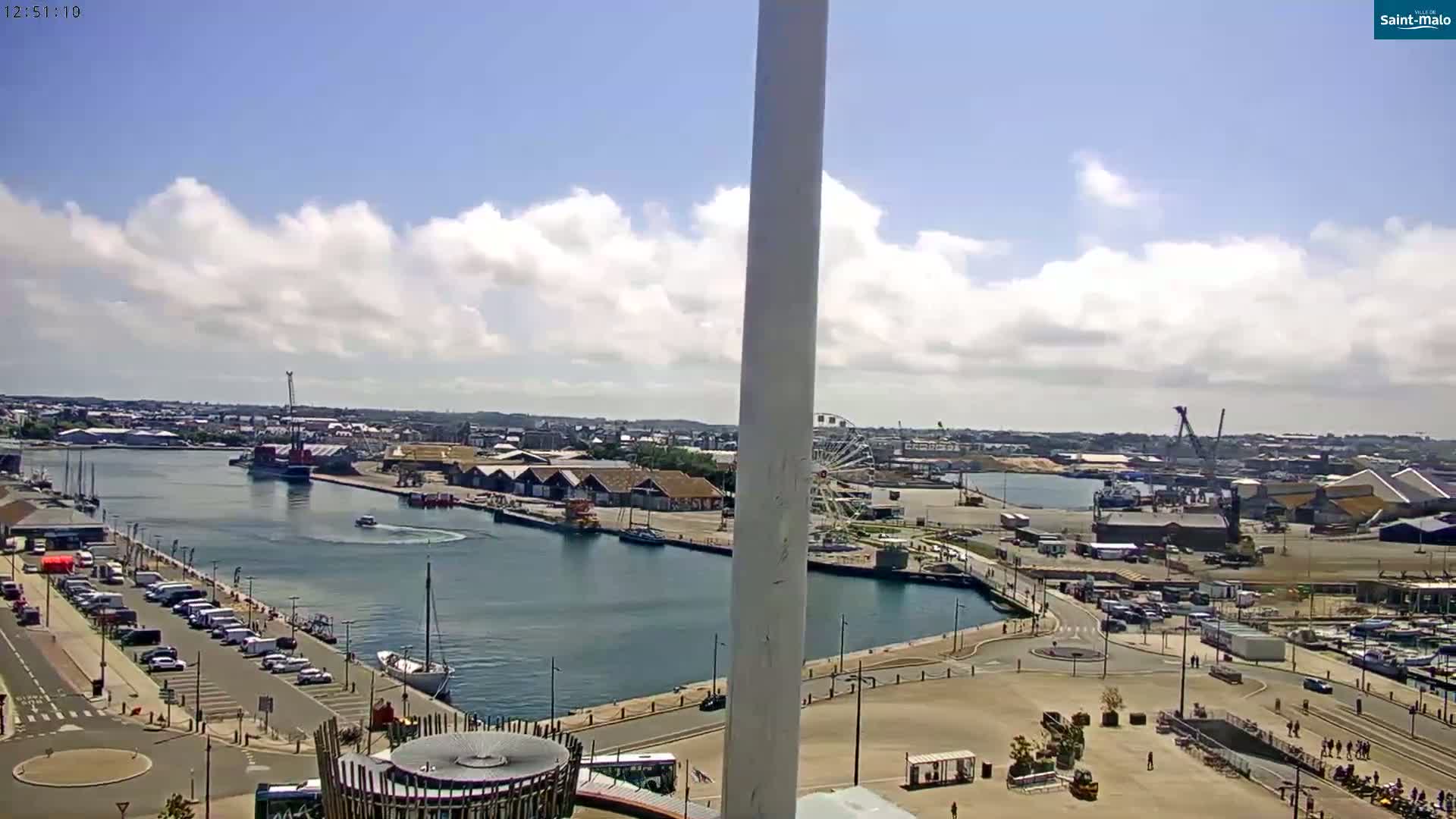 A partly cloudy day reveals a harbor scene with a marina, docked boats, a Ferris wheel, and parking lots, viewed from a high vantage point.