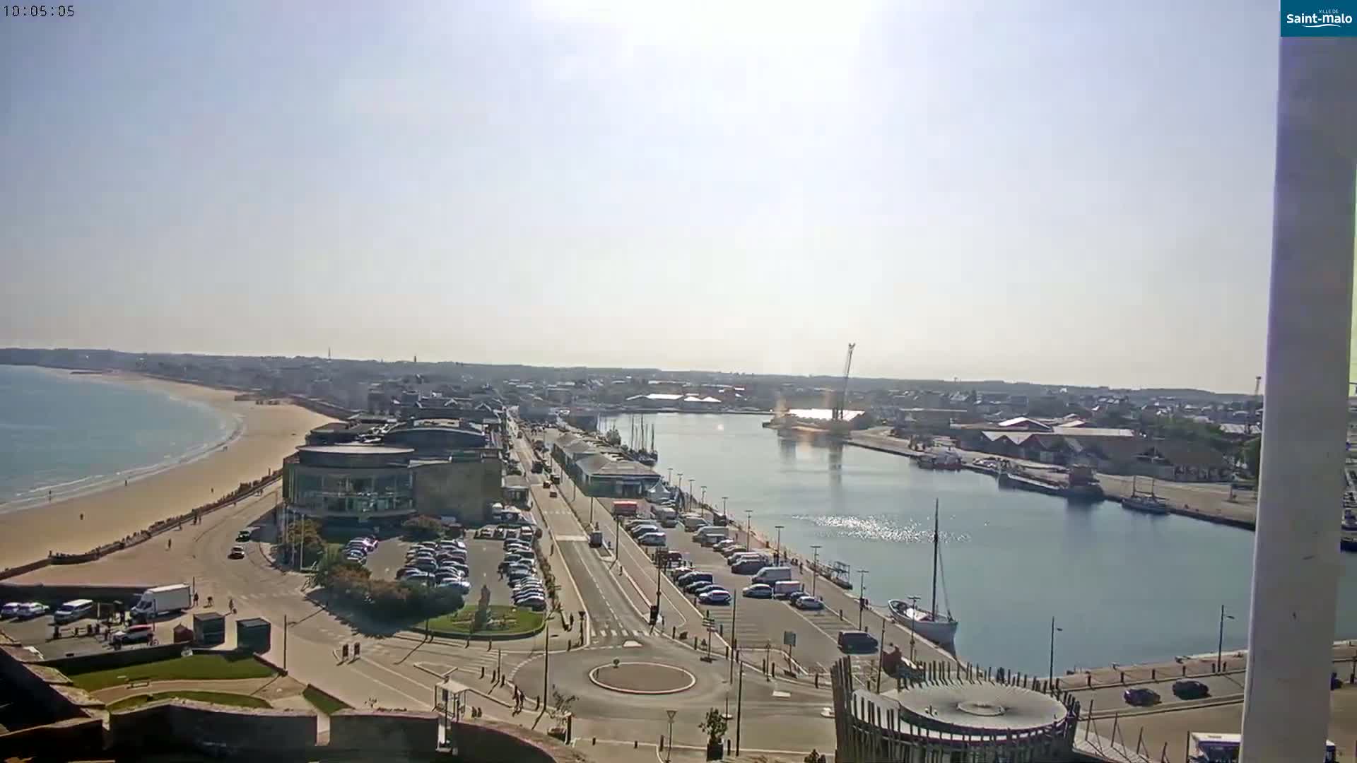 A sunny day overlooking a harbor town with a sandy beach, a calm waterway, and parked cars lining a road.