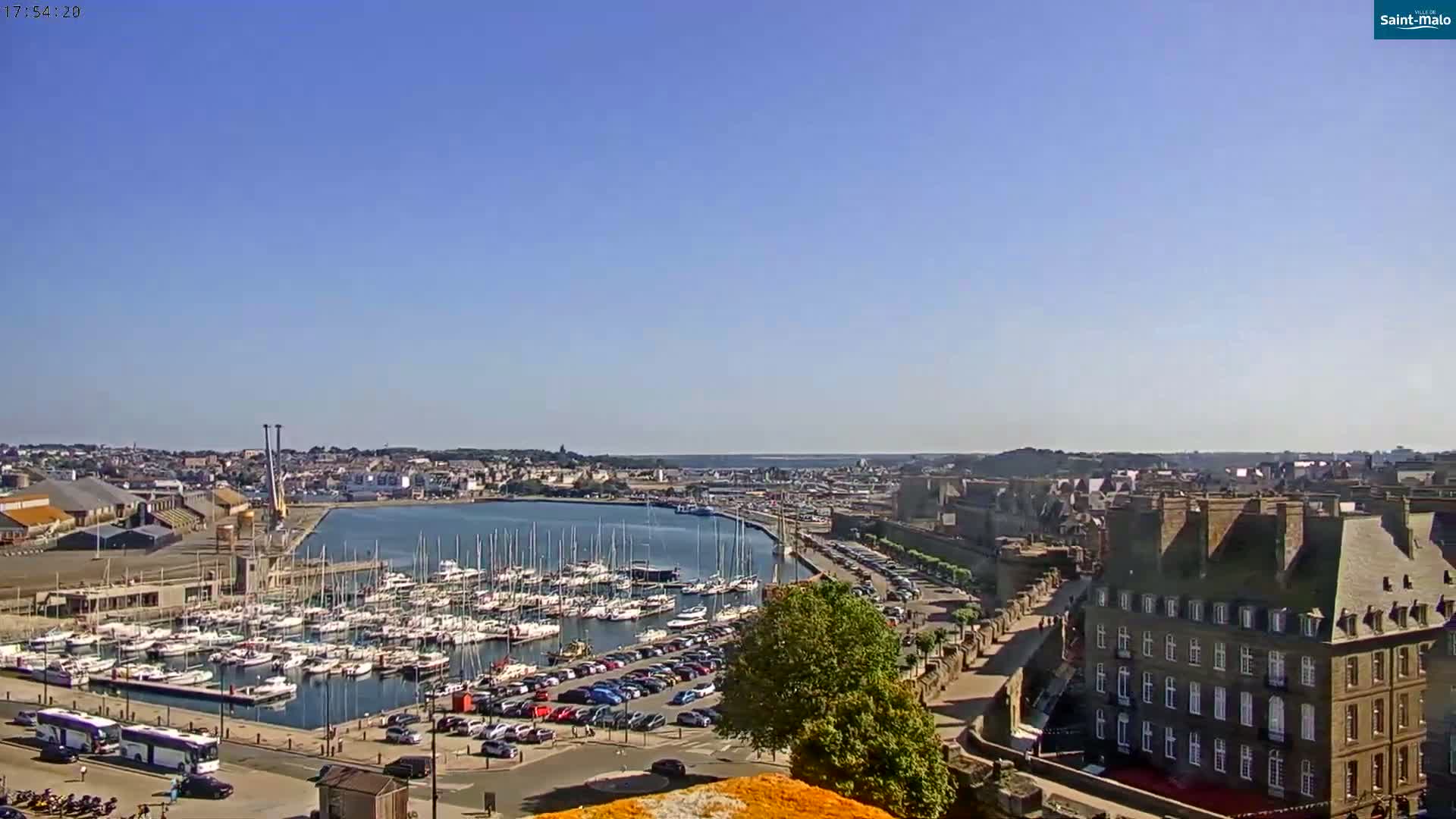 A harbor filled with sailboats is situated next to a city with stone buildings under a clear blue sky.