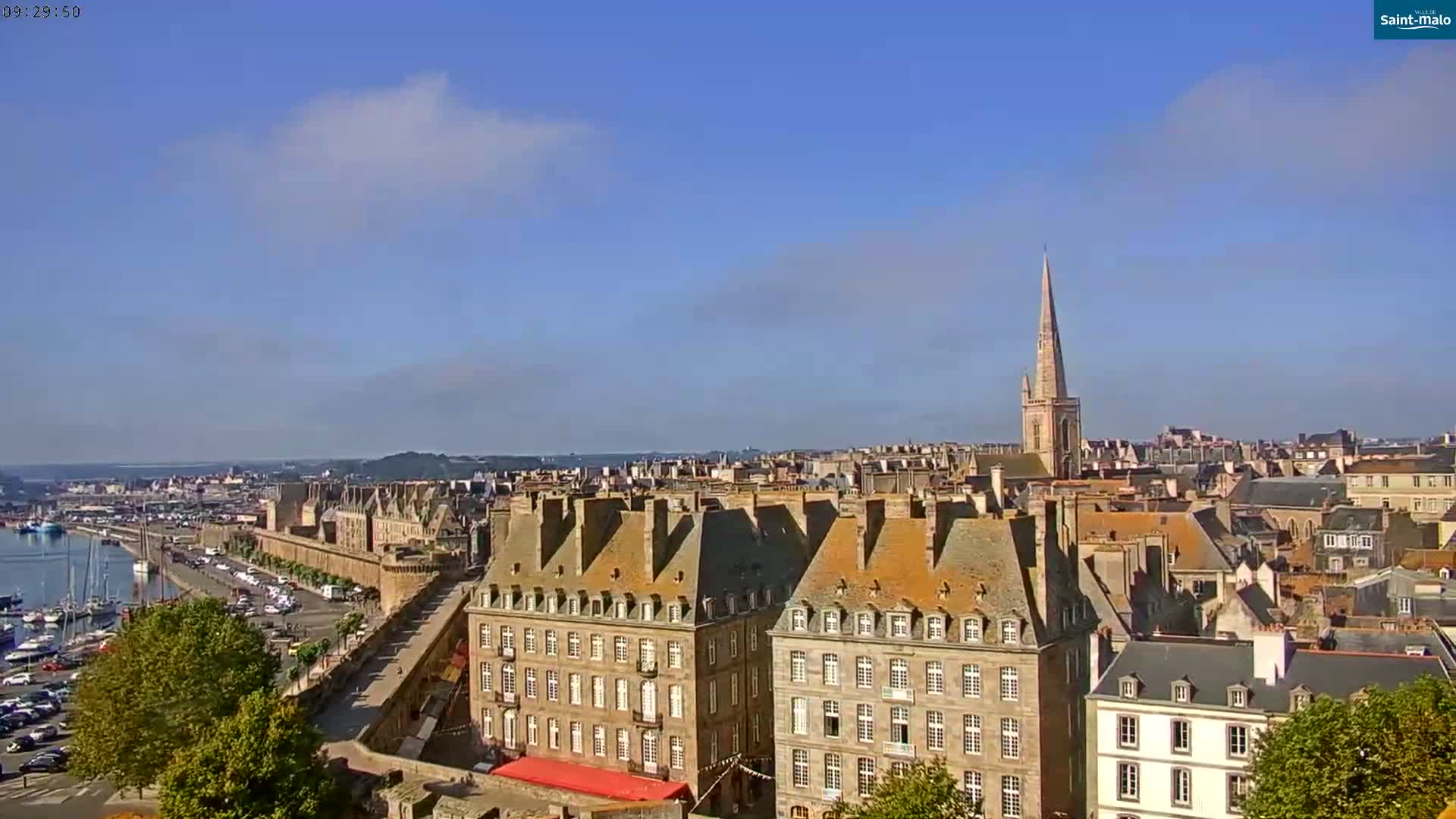 A partly sunny day reveals a panoramic view of a coastal city with a marina, stone buildings, and a tall church spire in the distance.
