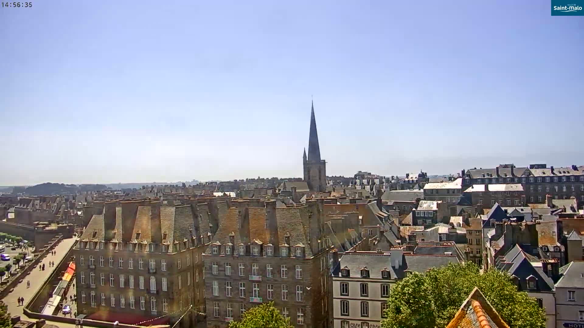 A high-angle, long shot on a clear, sunny day shows a cityscape of many buildings with dark gray slate roofs, a prominent church spire, and a glimpse of a walled area.