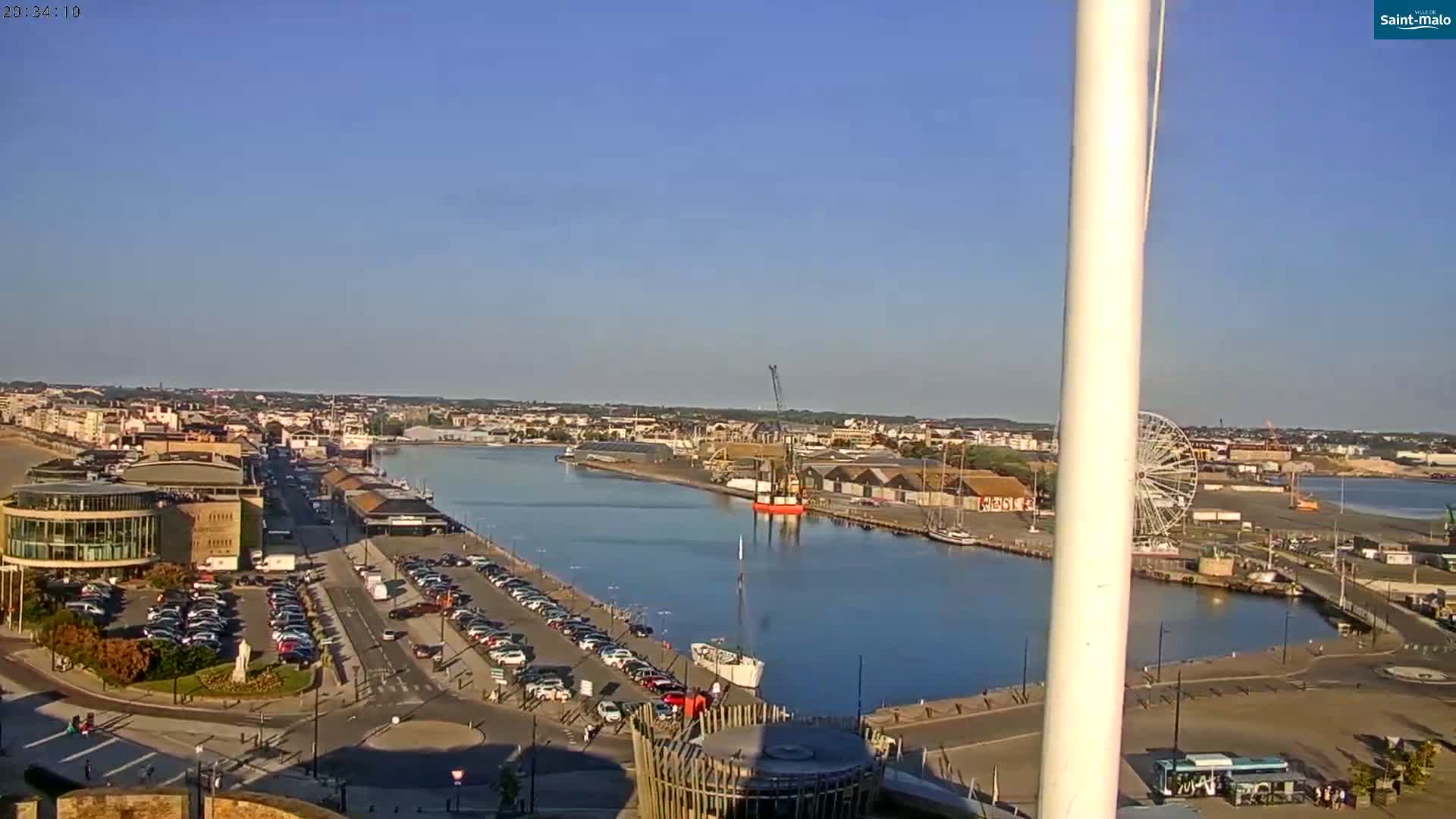 A harbor scene on a clear, sunny day shows a city, a body of water with boats, a Ferris wheel, and numerous parked cars.