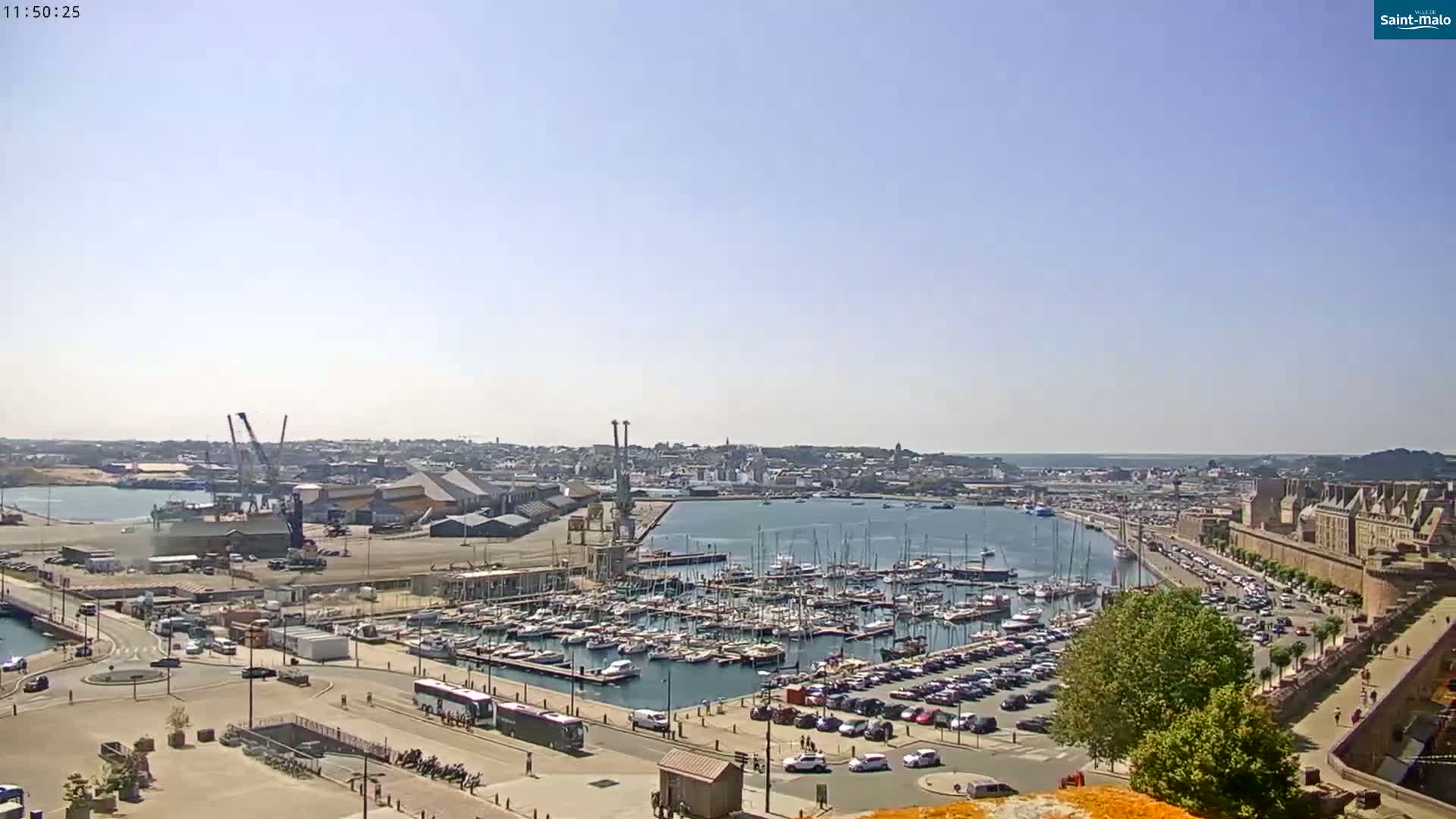 A sunny day overlooking a harbor filled with boats, adjacent to a city with a large stone wall and parking lots.