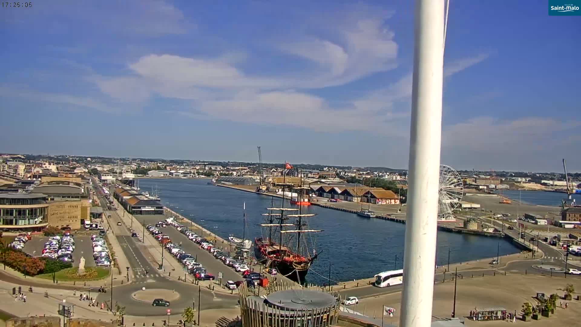 A mostly sunny day reveals a harbor scene with a large, black-sailed ship docked near a Ferris wheel, numerous parked cars, and buildings lining the waterfront.