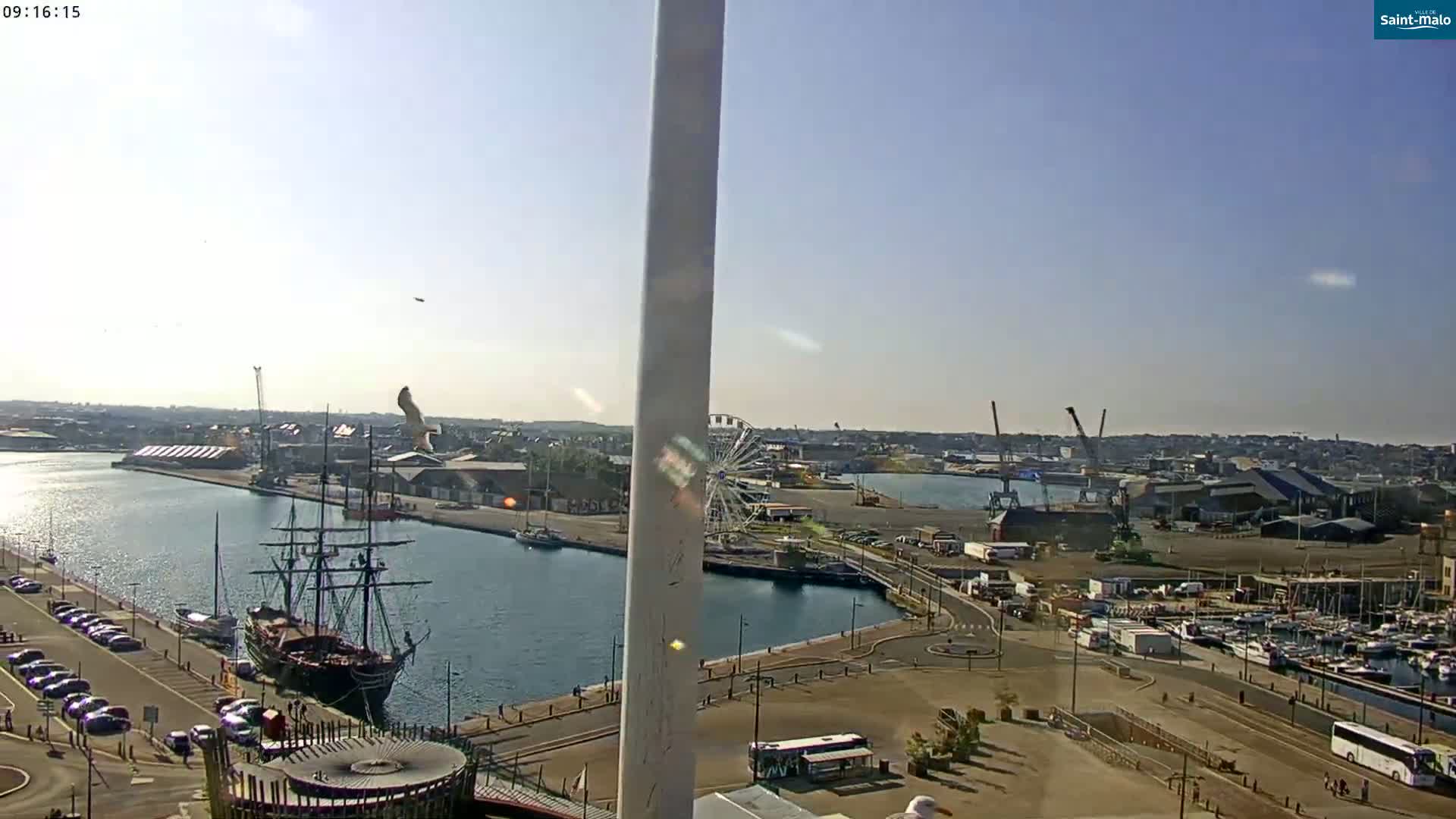A sunny harbor scene with a large sailing ship, a Ferris wheel, and numerous boats docked at a marina.