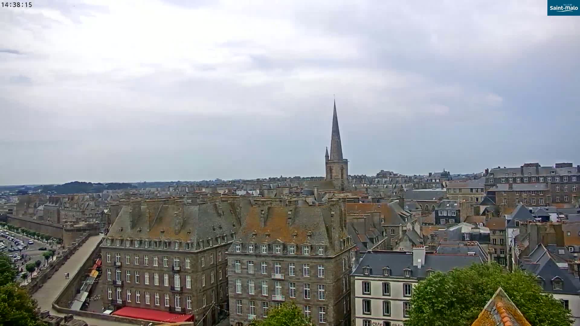 A panoramic view of a European city under a cloudy sky shows many stone buildings with steeply pitched roofs, including a tall church spire.