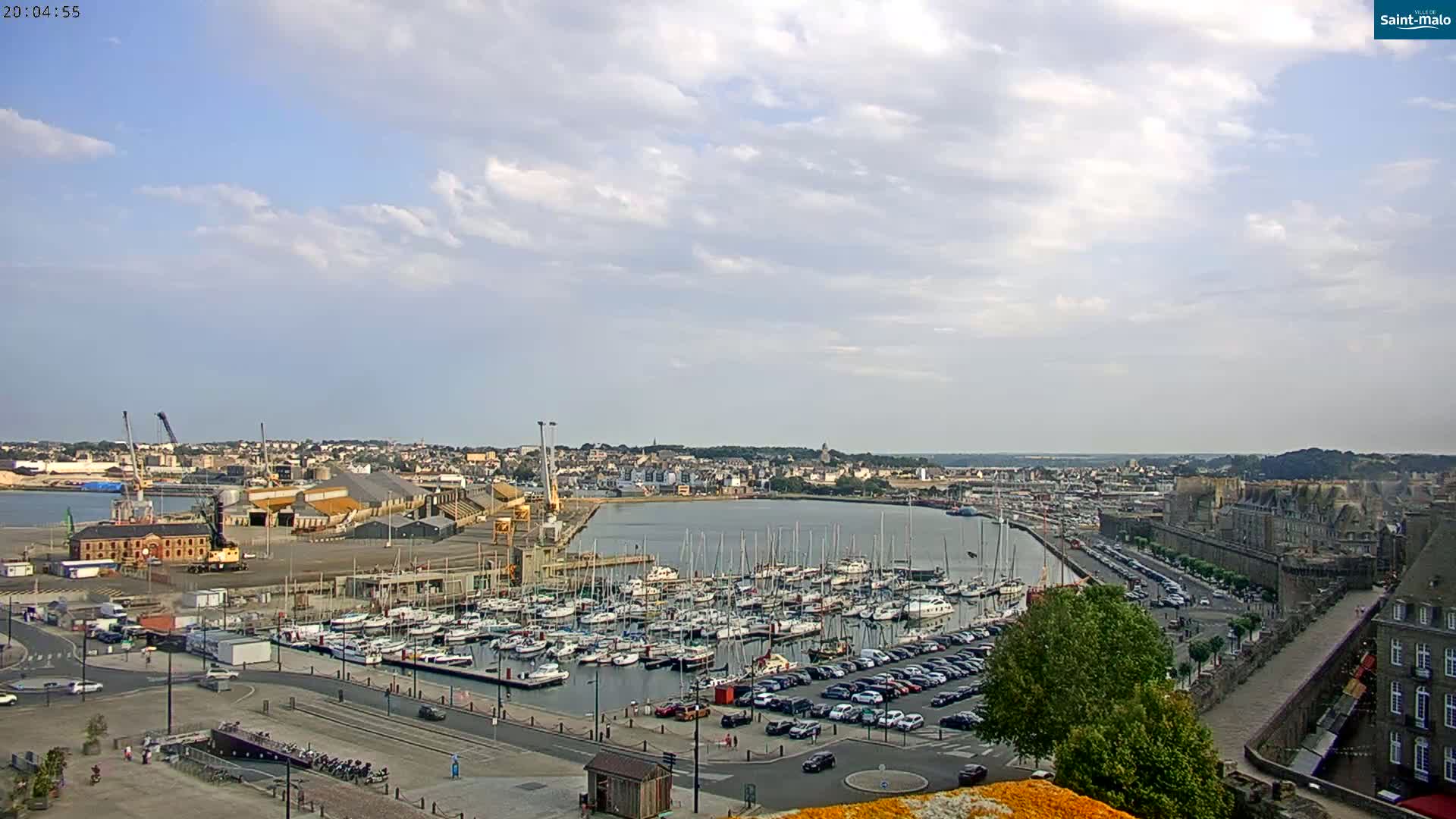 A partly cloudy day reveals a harbor filled with boats, a city, and a stone wall, with cars parked nearby.