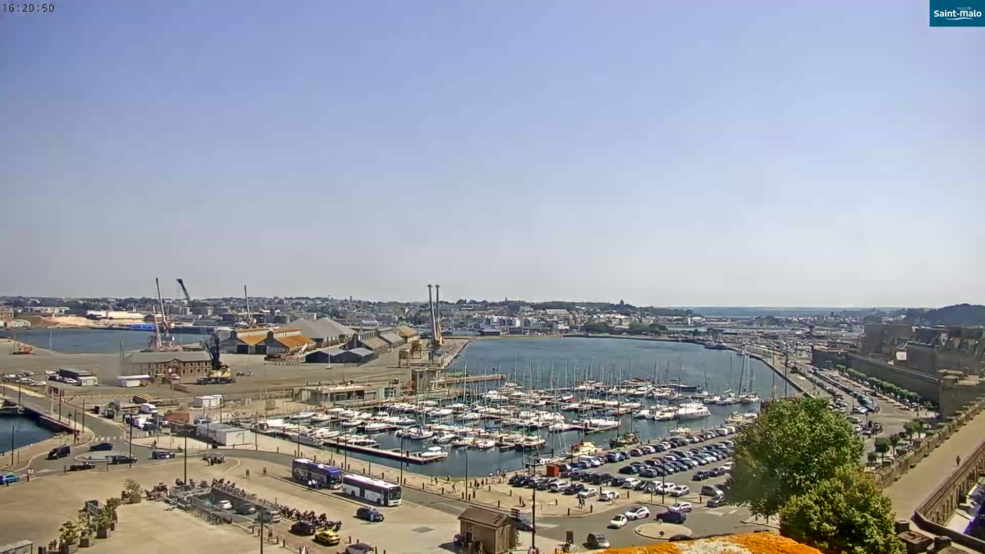 A harbor filled with boats under a clear blue sky, with a city and surrounding landscape visible in the distance.