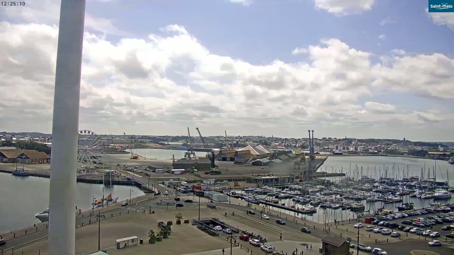 A partly cloudy day reveals a harbor filled with sailboats and motorboats, industrial areas, and a Ferris wheel in the distance.