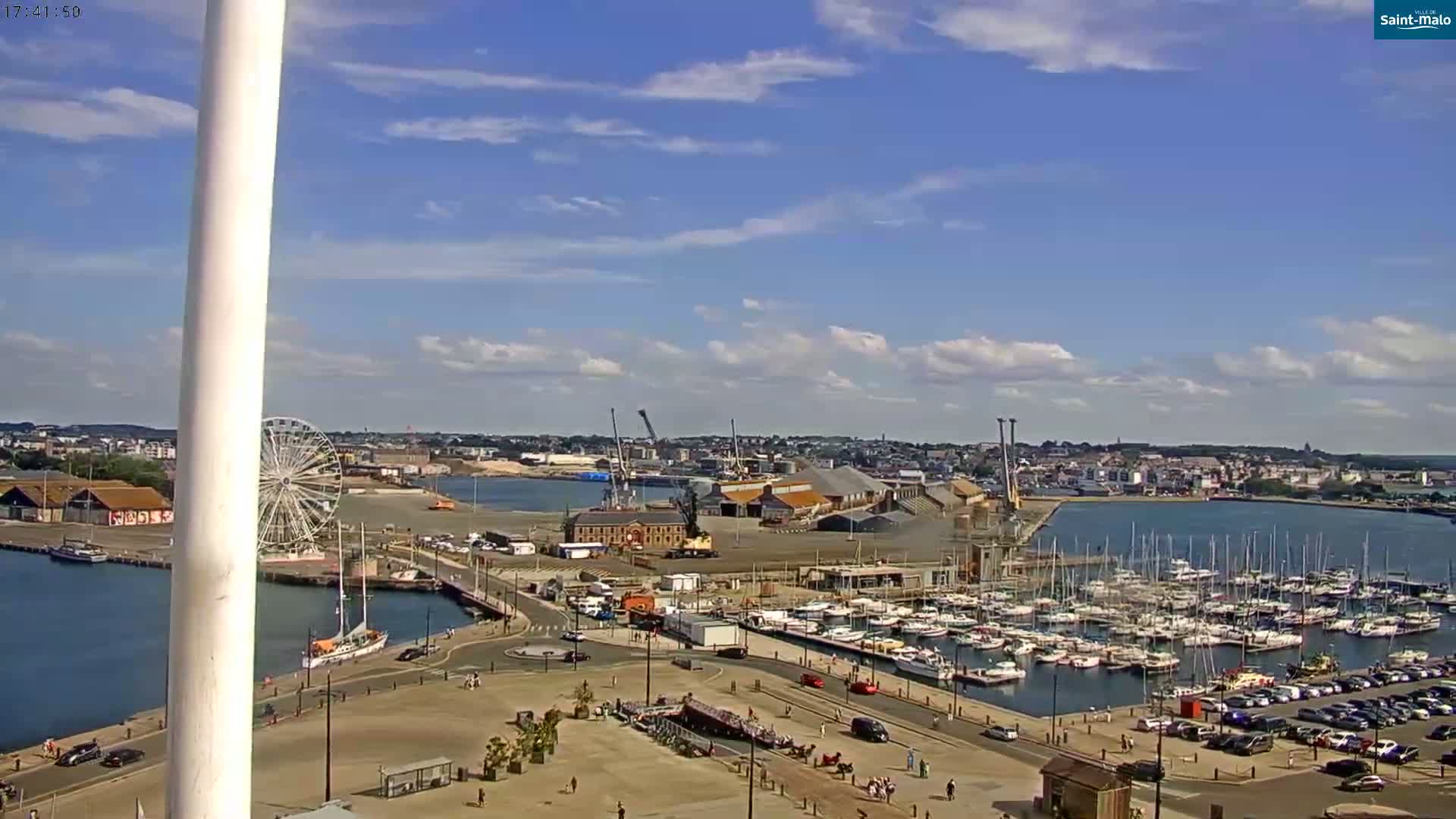 A harbor filled with sailboats under a partly cloudy sky, next to a Ferris wheel and a town.
