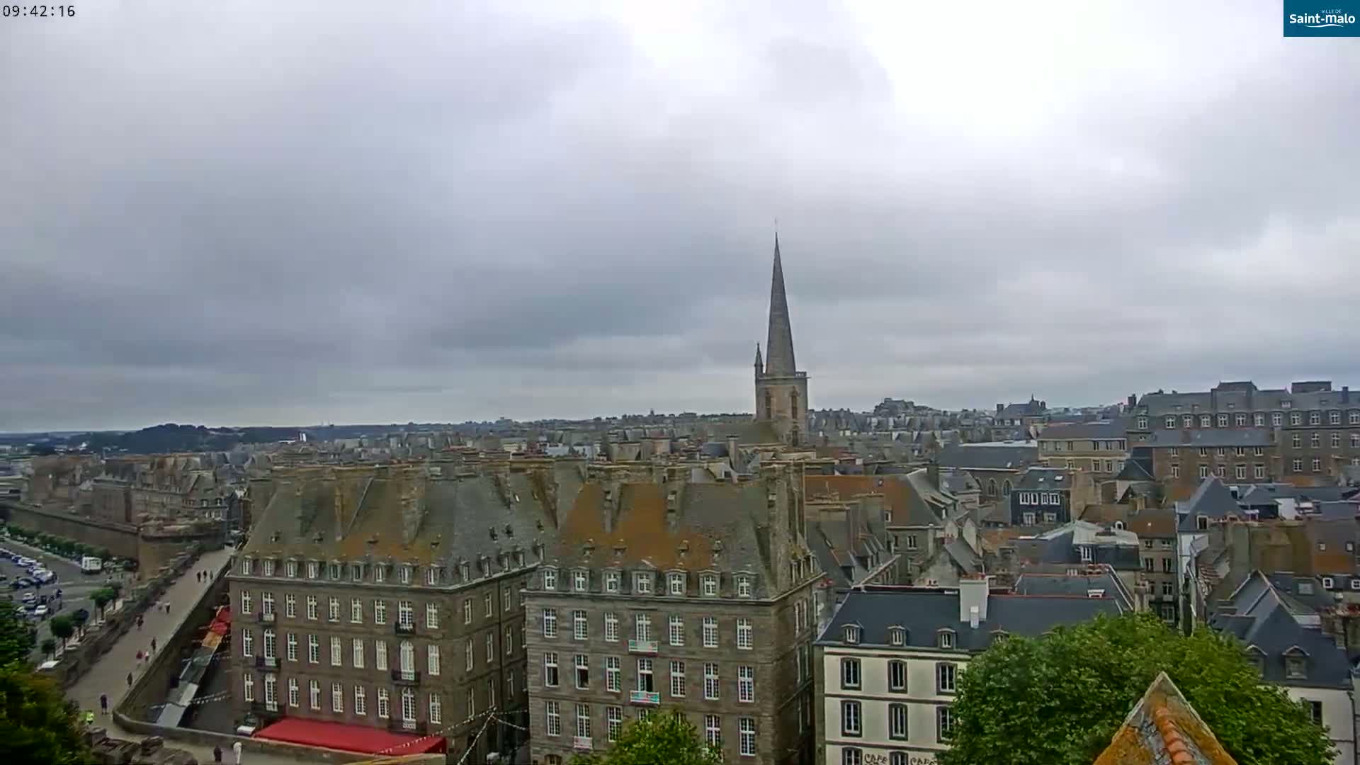 A panoramic view of a European city under a cloudy sky shows many buildings with grey slate roofs, a tall church spire, and a walled area.