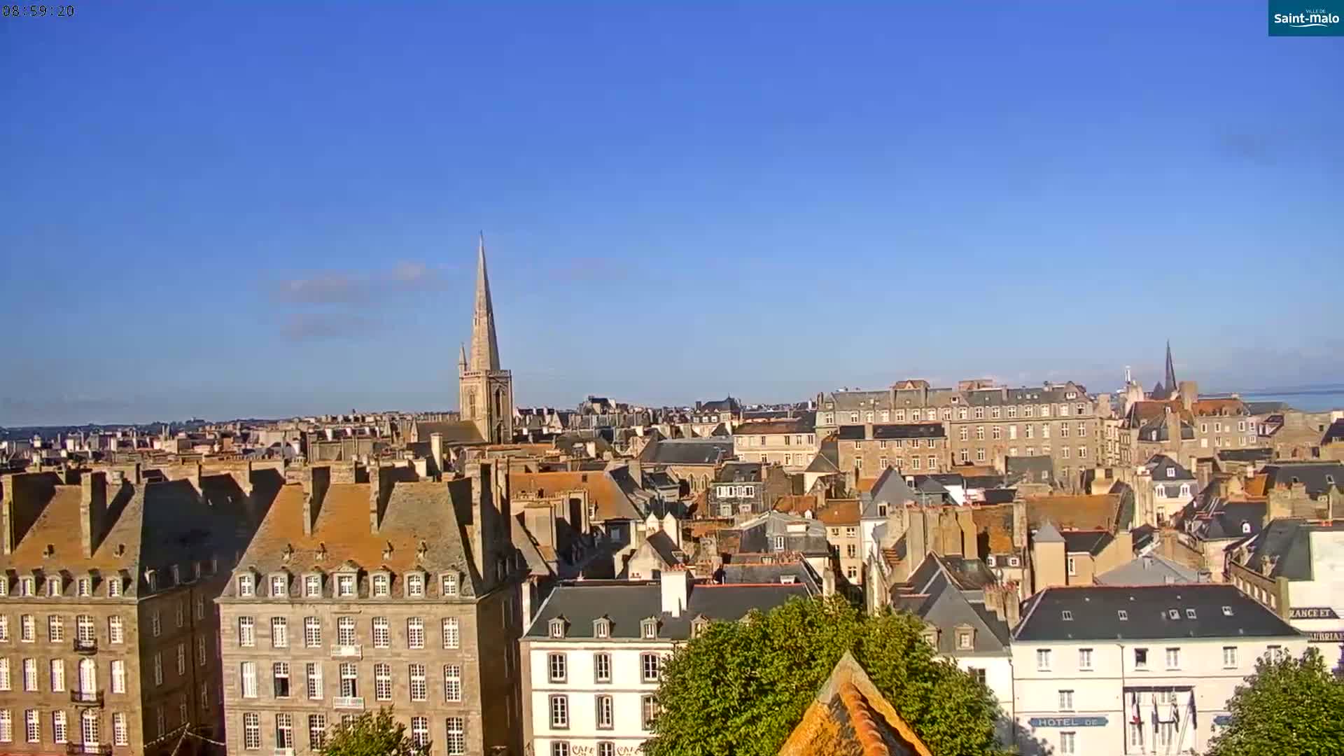 A panoramic view of a city with many tightly packed buildings under a clear blue sky, showcasing a prominent church steeple in the mid-ground.