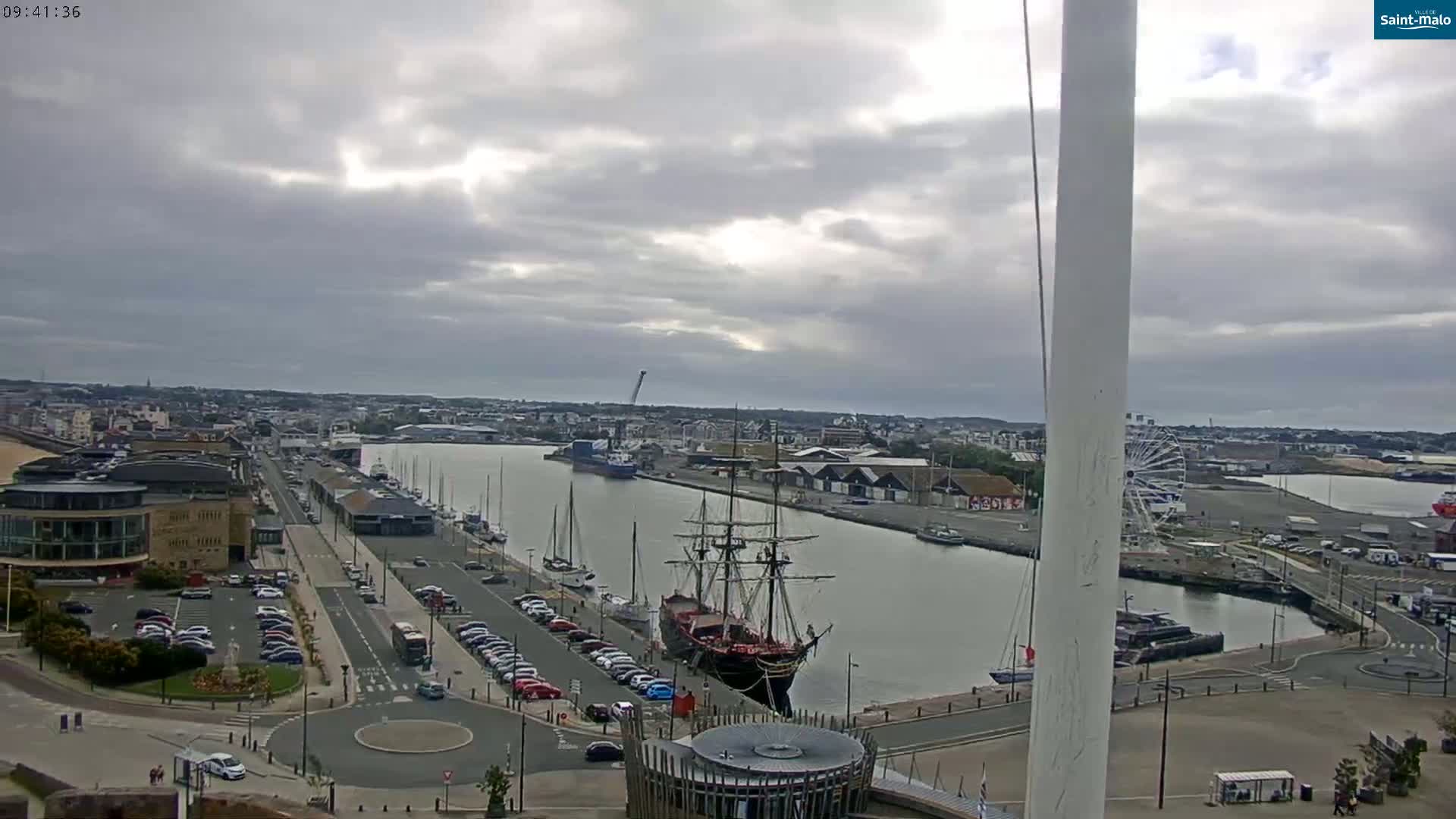 A mostly cloudy day reveals a harbor scene with a large sailing ship docked near a waterfront with parked cars and buildings,  a Ferris wheel visible in the distance.