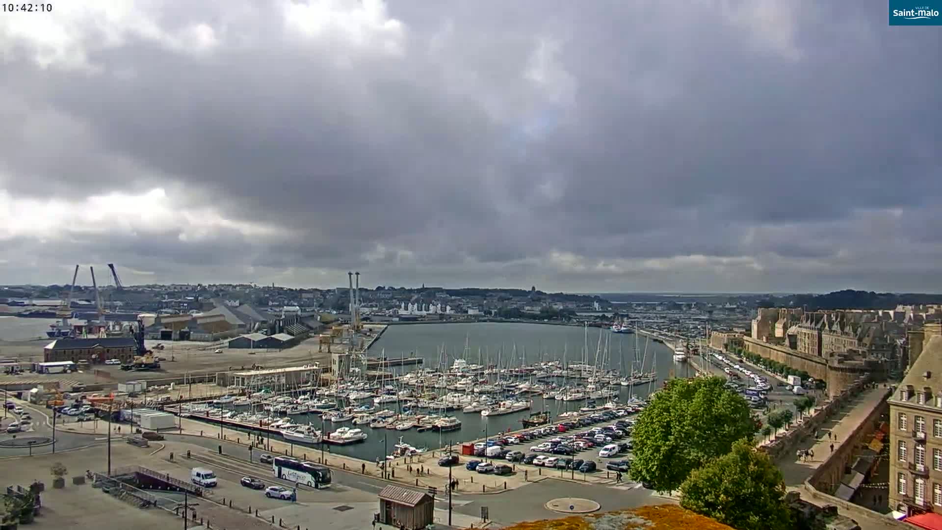 A harbor filled with numerous sailboats and yachts is situated under a mostly cloudy sky, next to a city with ancient-looking walls and buildings.