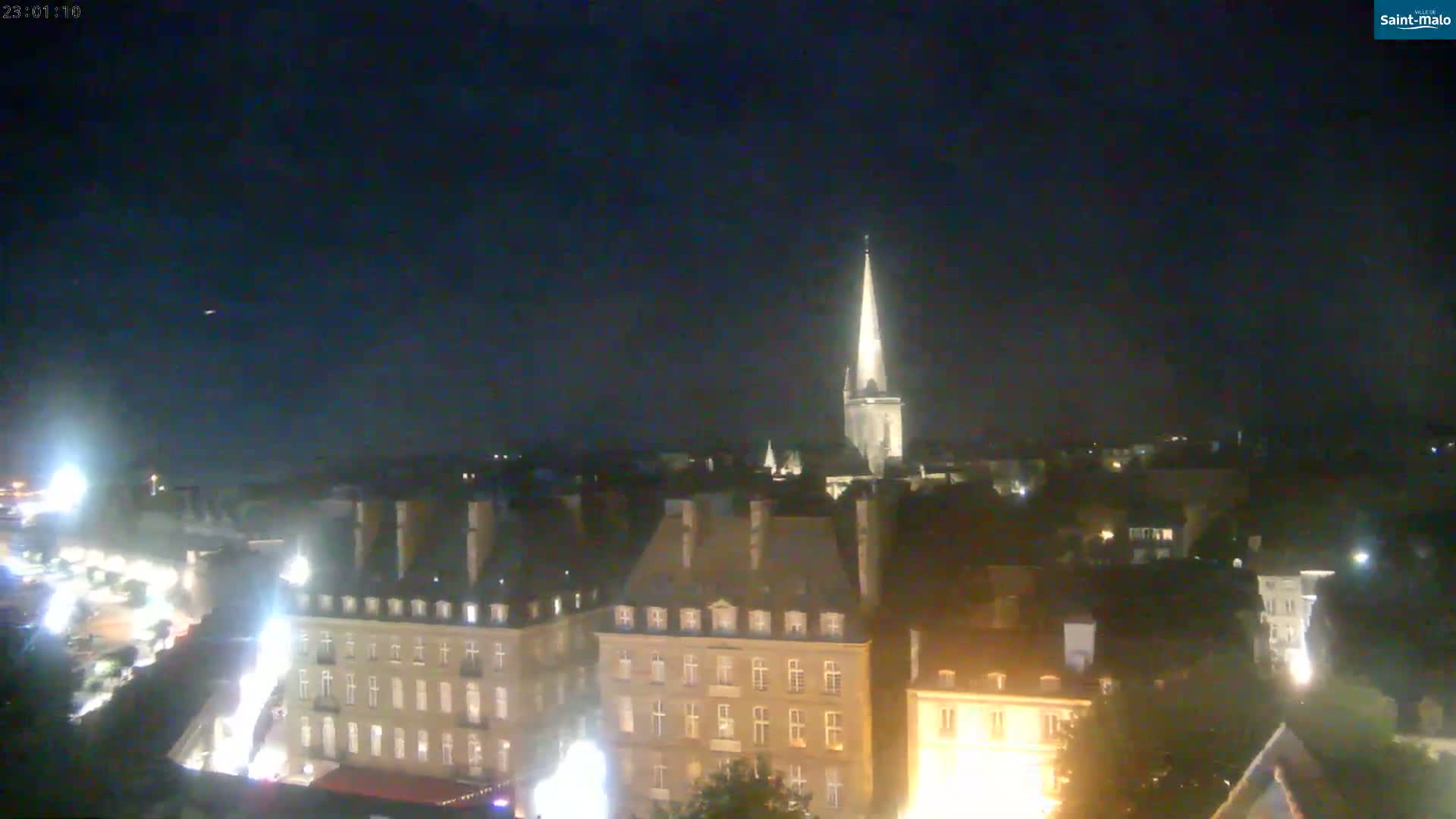 A nighttime, aerial view shows a city's illuminated buildings and a tall church spire under a clear, dark sky.