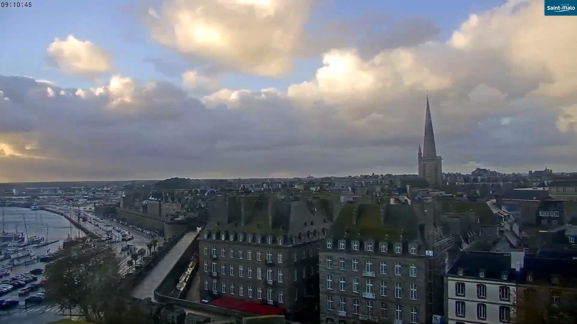 A panoramic view of a historic coastal city features a busy marina with boats, dense stone buildings, and a prominent church spire, all under a partly cloudy sky where sunlight breaks through dark clouds onto damp streets.