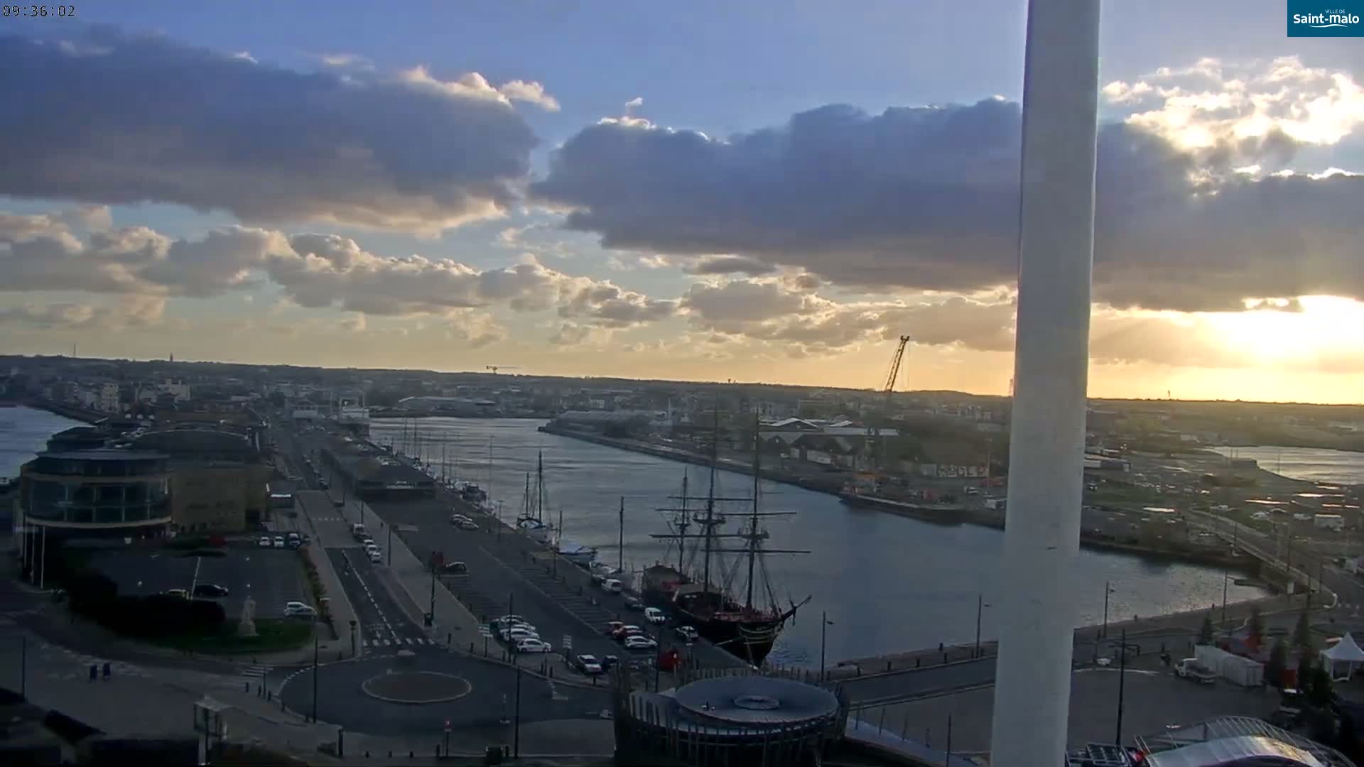 An elevated panoramic view captures a port city at either sunrise or sunset, featuring a calm waterway bordered by roads with traffic and various buildings, including a large historic sailing ship docked alongside, under a partly cloudy sky with golden sunlight breaking through.