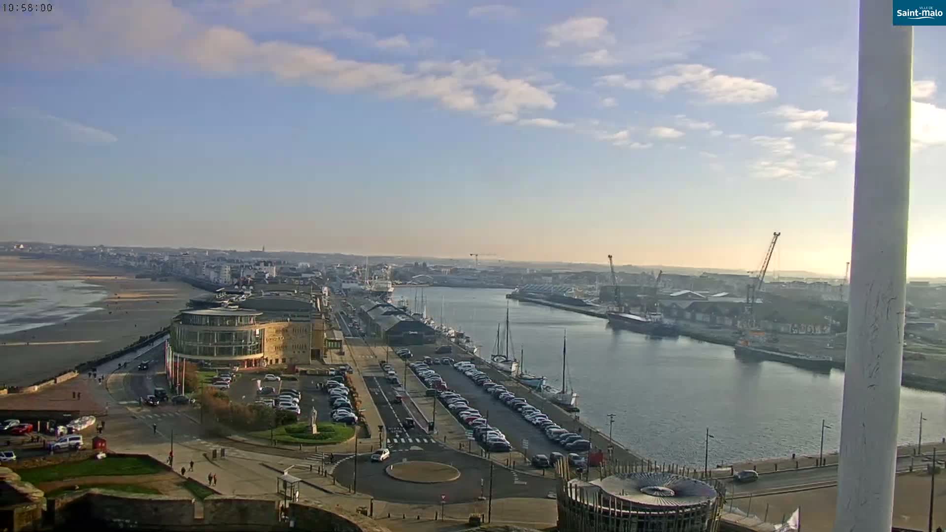 An elevated panoramic view captures a port city at either sunrise or sunset, featuring a calm waterway bordered by roads with traffic and various buildings, including a large historic sailing ship docked alongside, under a partly cloudy sky with golden sunlight breaking through.