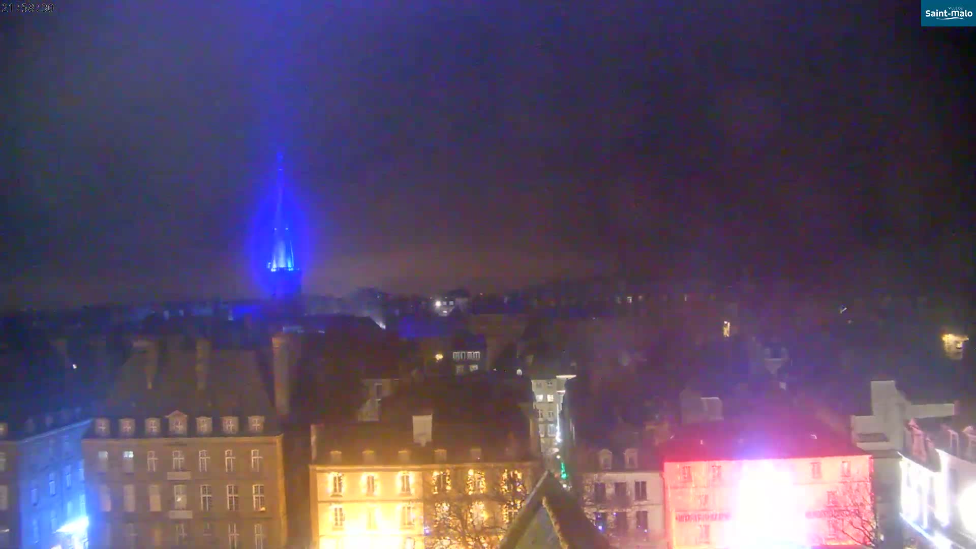 An elevated panoramic view captures a port city at either sunrise or sunset, featuring a calm waterway bordered by roads with traffic and various buildings, including a large historic sailing ship docked alongside, under a partly cloudy sky with golden sunlight breaking through.