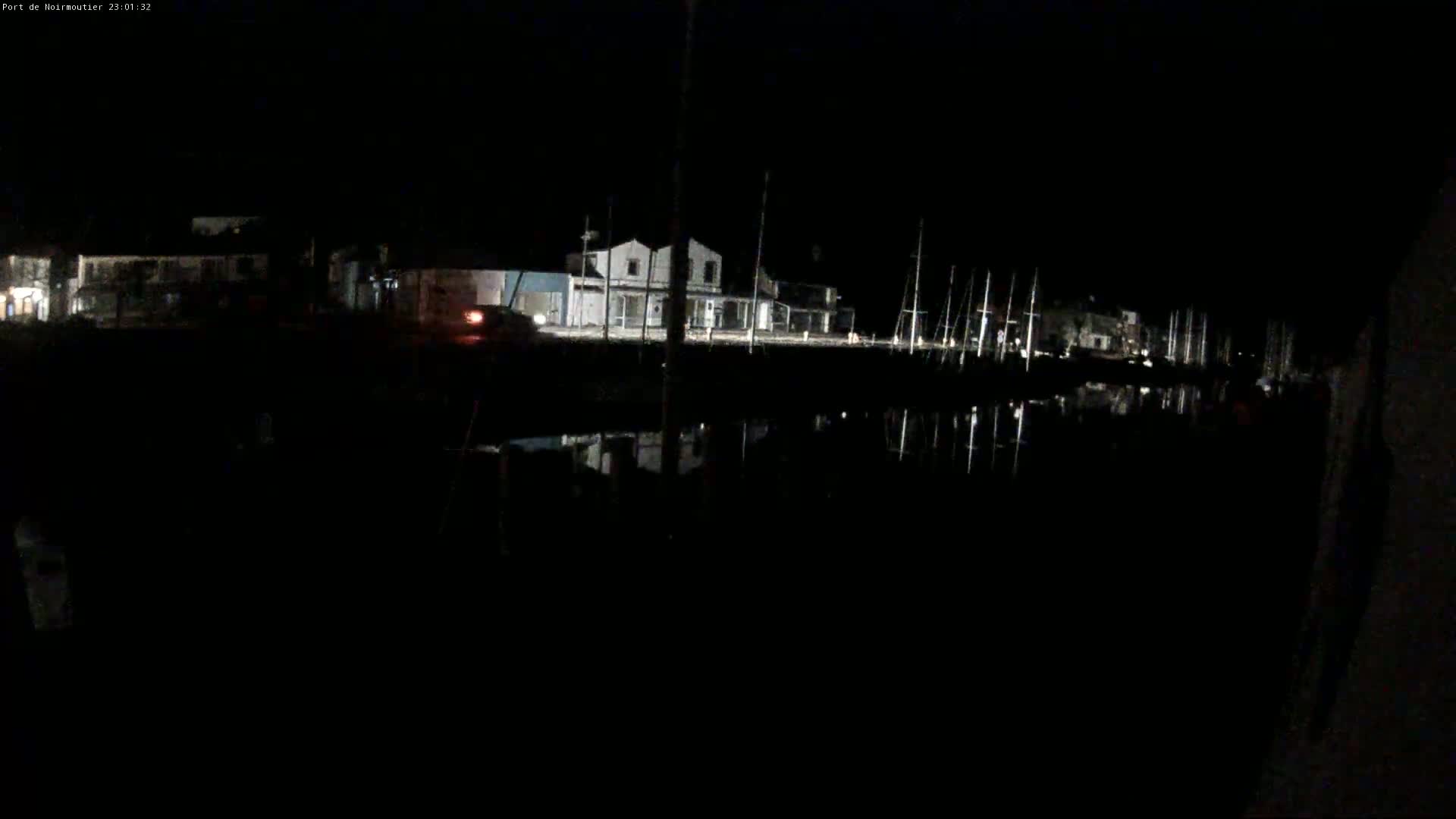 At night, a dark body of water reflects the lights of buildings and several masts lining the opposite shore.