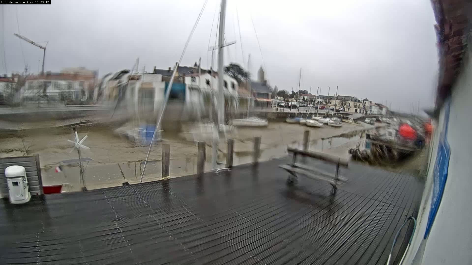 A wet, overcast day at a harbor shows numerous boats moored at a dock, with a muddy shoreline and a bench visible on the dock.