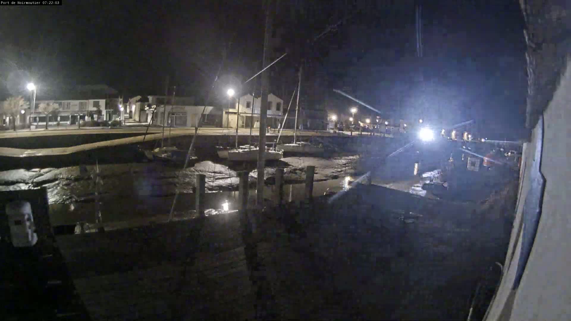 A nighttime view of a harbor with several sailboats docked at low tide, under partly cloudy skies.