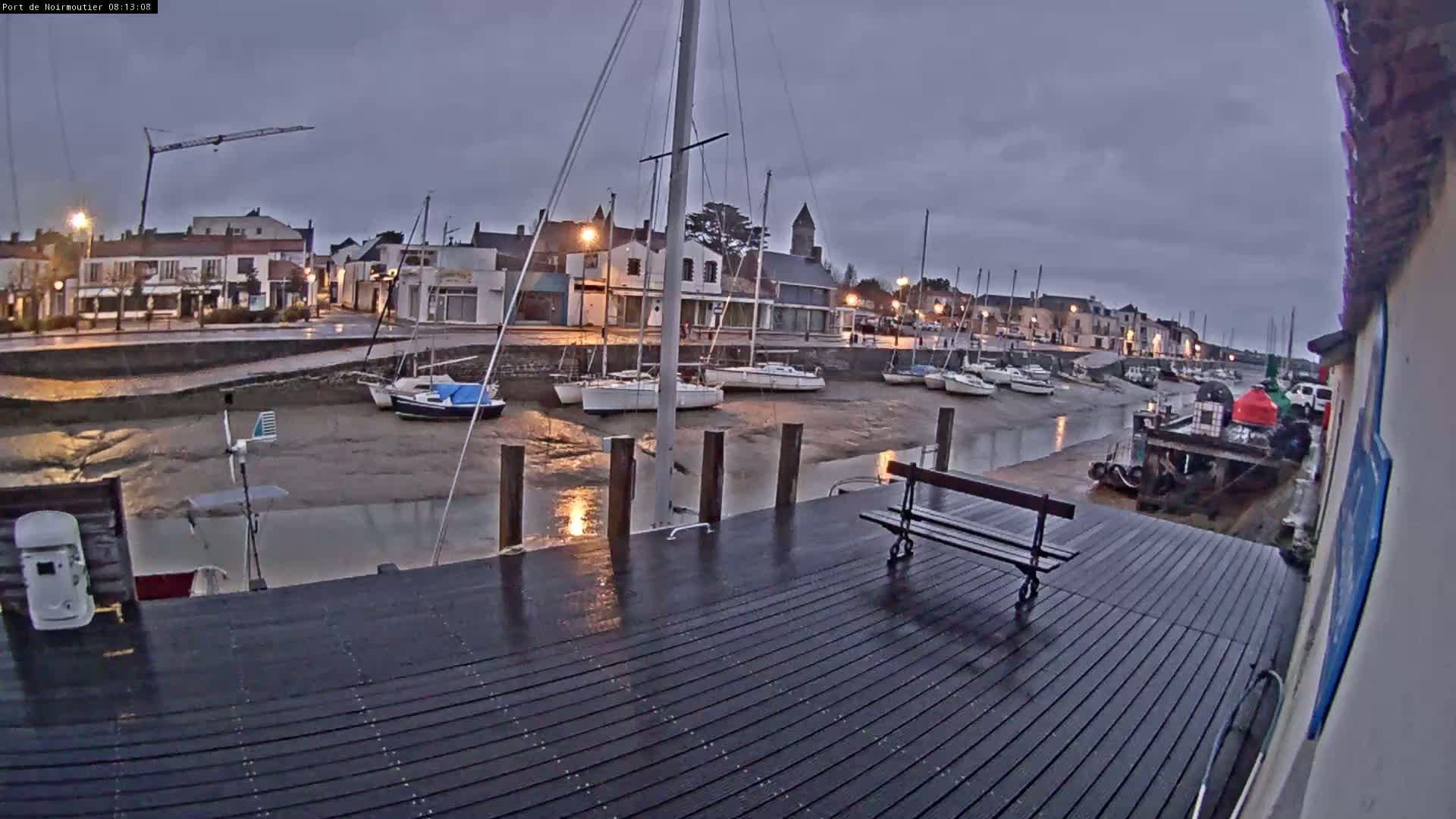 A wet wooden dock overlooks a harbor at low tide, where several sailboats are moored near a small town under an overcast sky.
