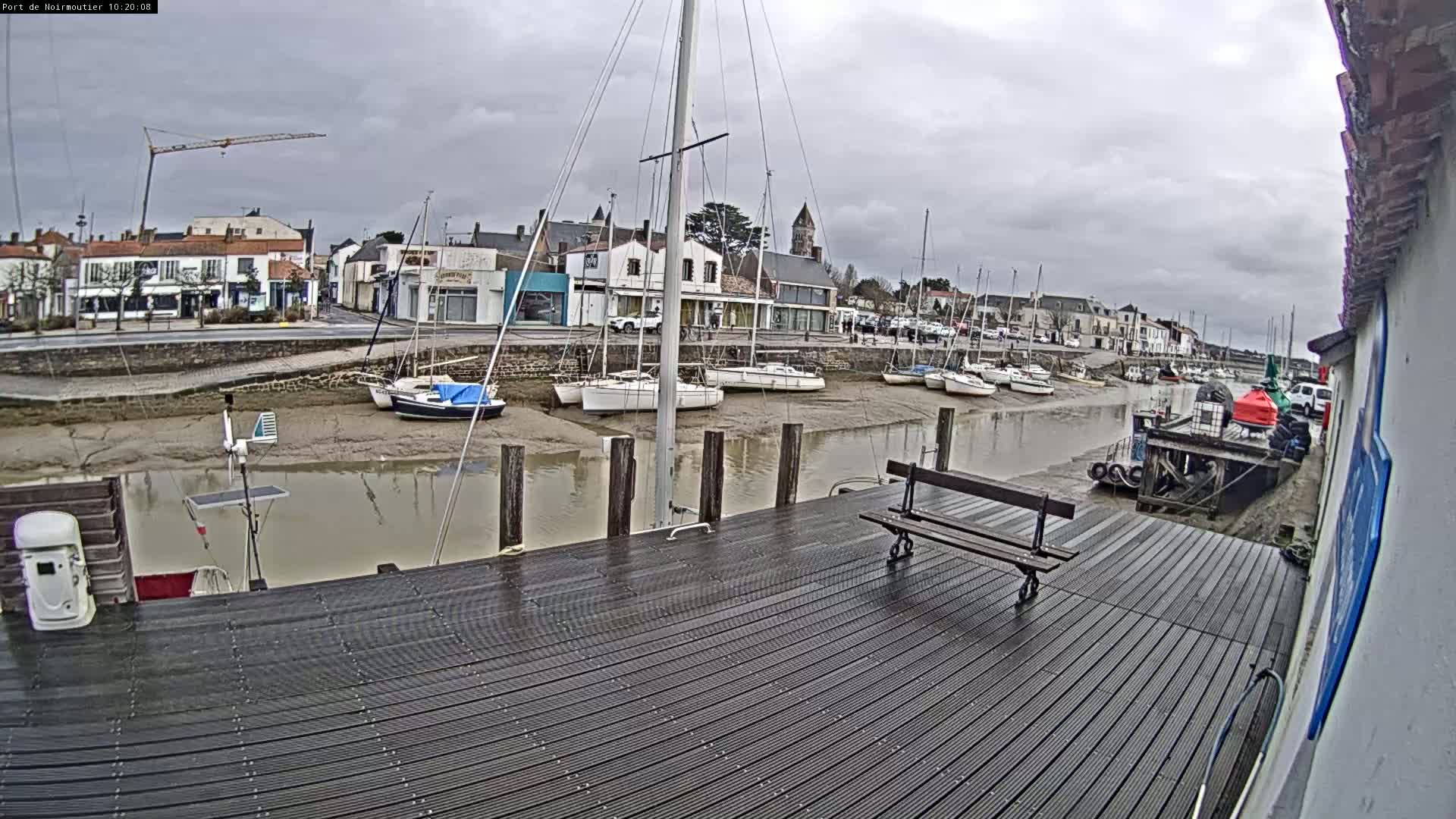A dark-colored wooden dock sits overlooking a calm harbor with several sailboats at low tide under a cloudy sky.