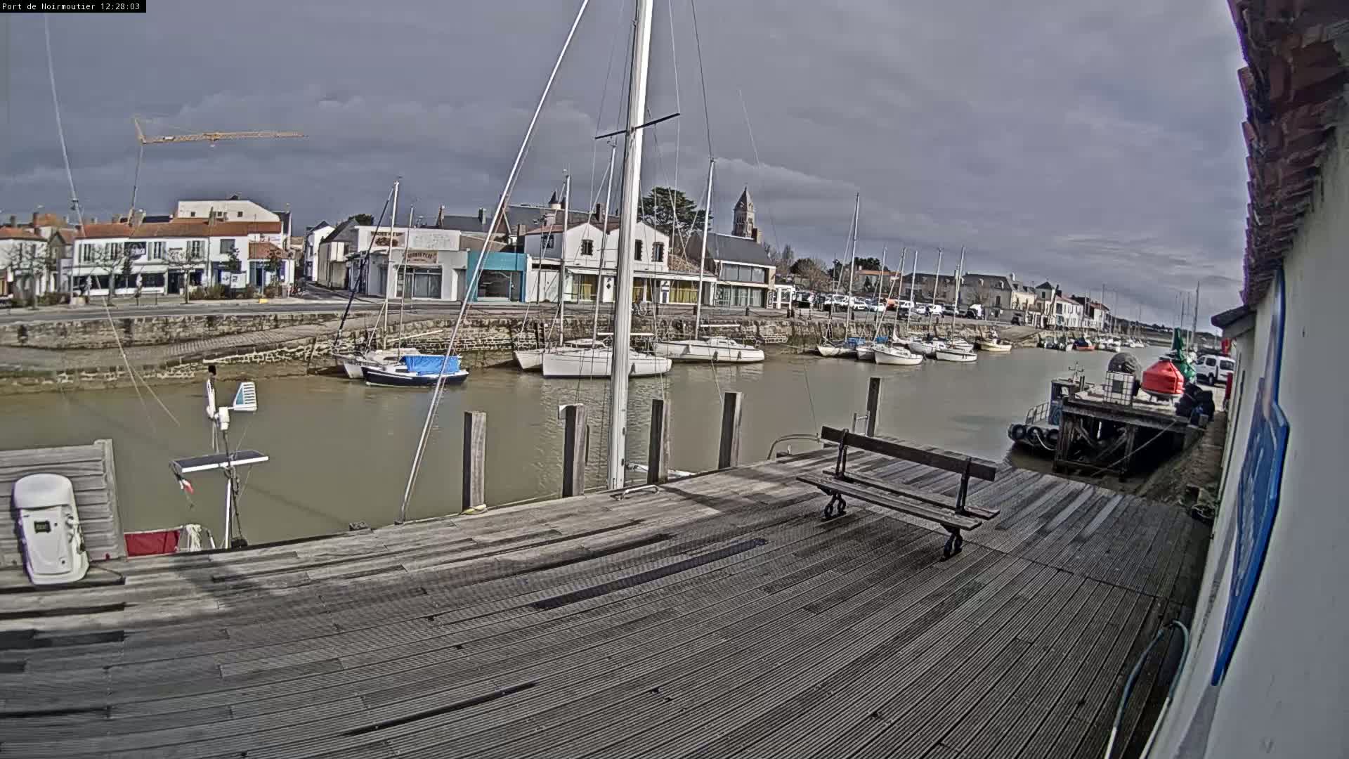 A mostly cloudy day reveals a harbor with numerous sailboats moored along a wooden dock, a small town in the background.
