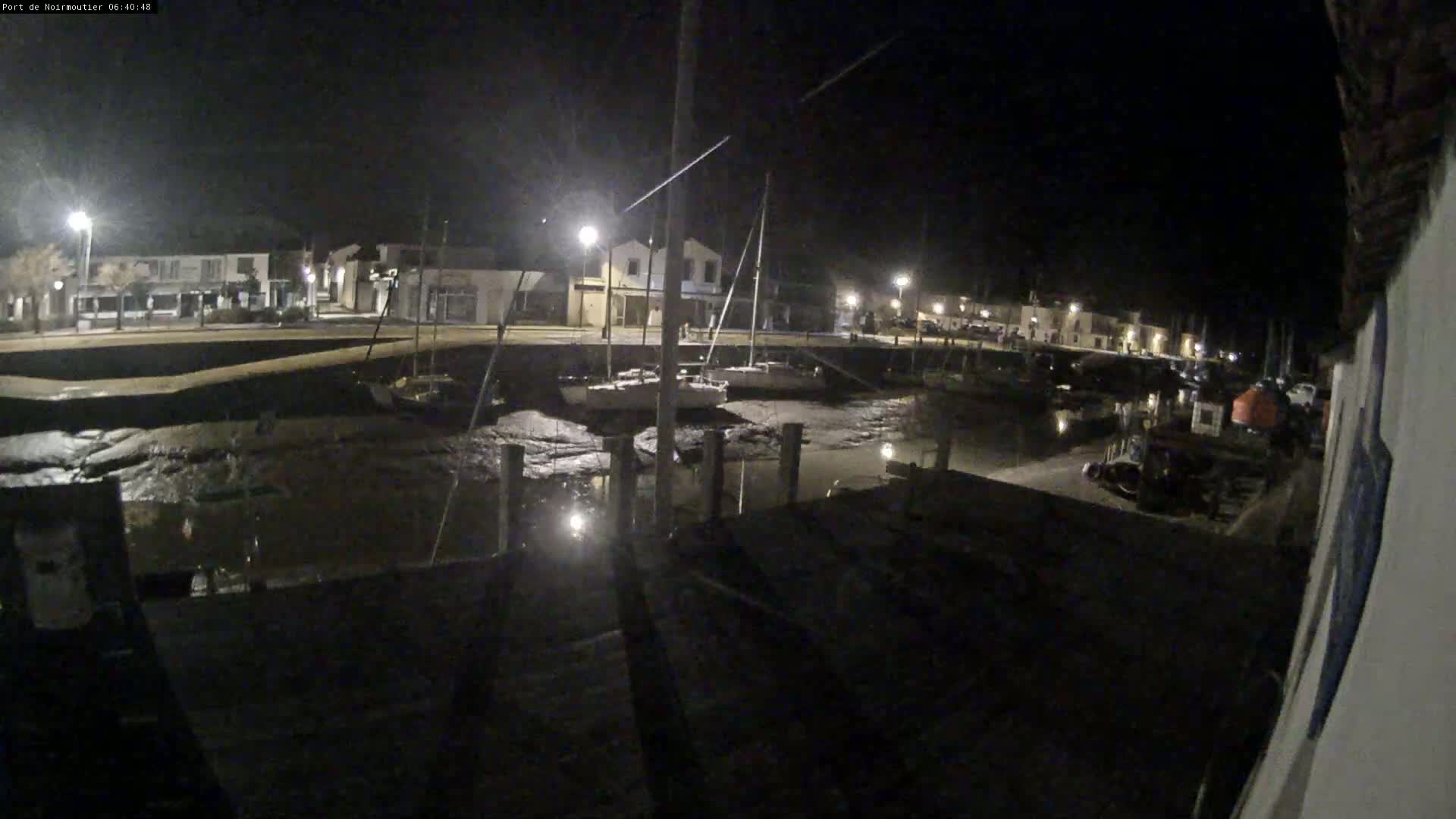 At night, several sailboats are docked in a calm harbor with exposed mudflats visible at low tide, under artificial light.