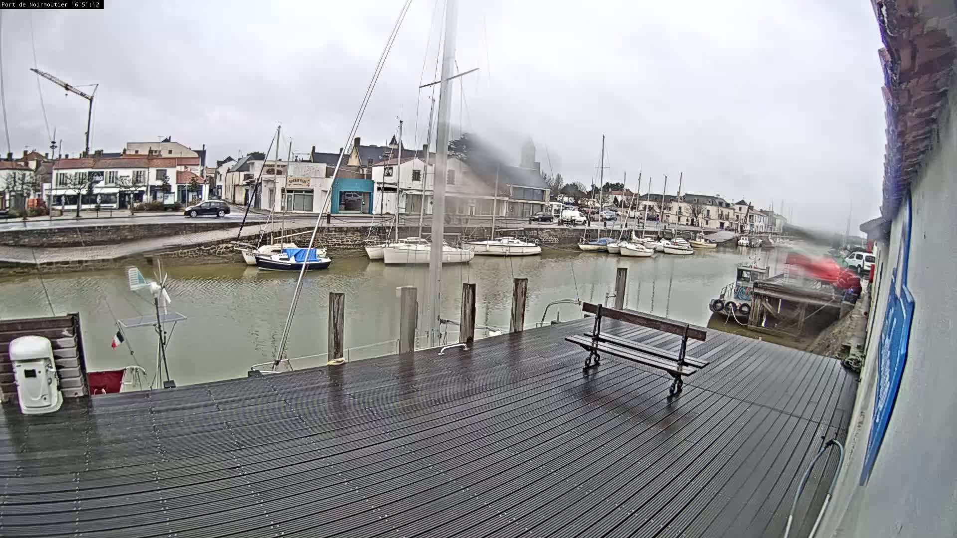 A wet dockside with several sailboats moored in calm water, under an overcast sky, in front of a town.