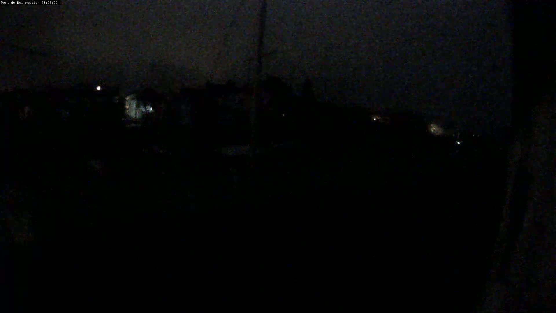 A nighttime, dark outdoor scene shows faintly lit buildings and a few distant lights under a cloudy sky.
