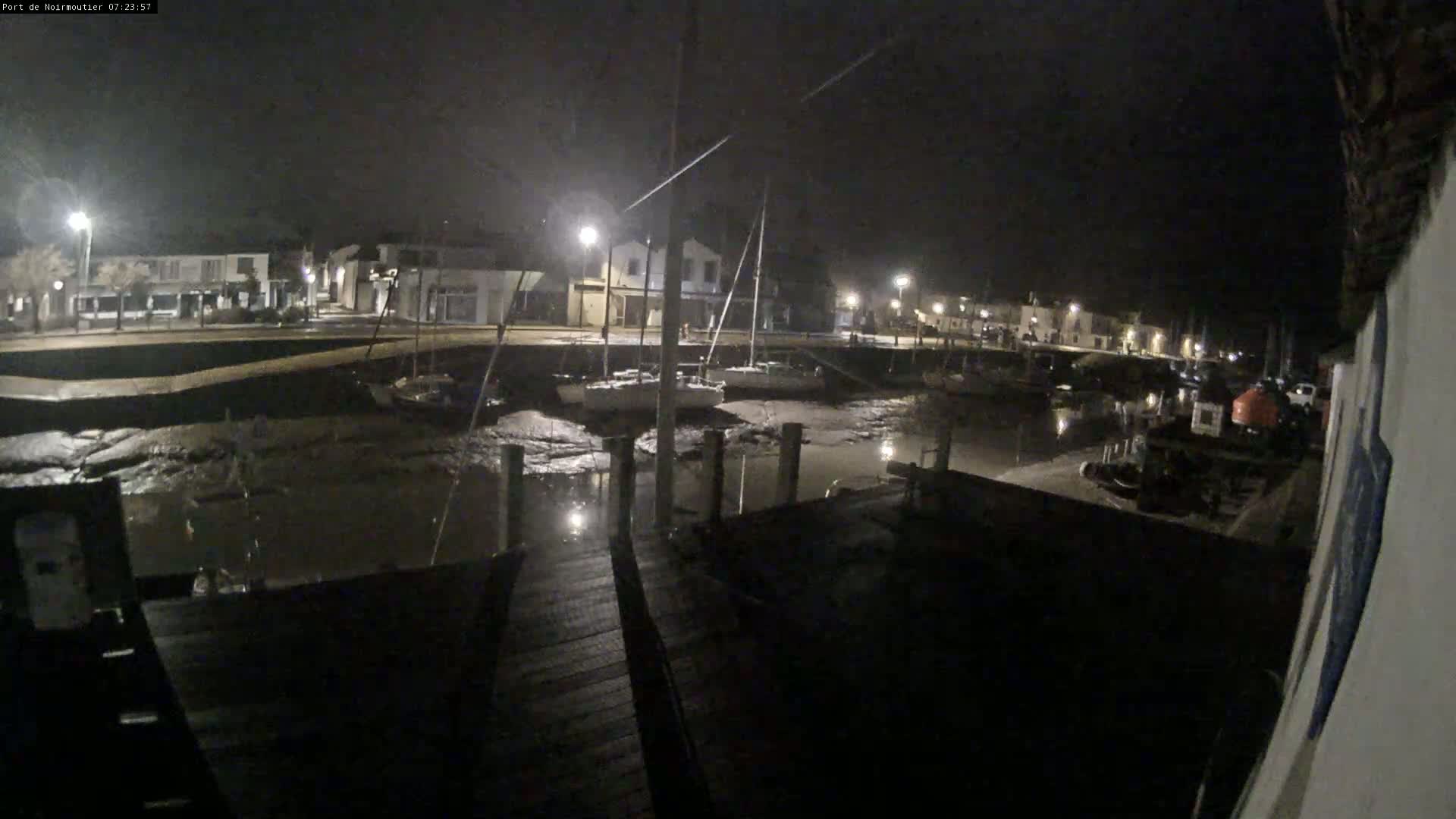 A nighttime view of a harbor with boats docked at low tide, illuminated by streetlights, under a dark sky.