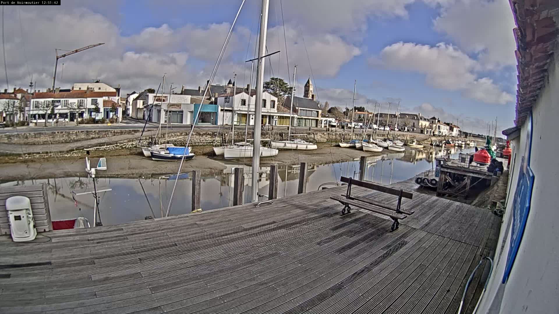 A mostly sunny day reveals a waterfront town with sailboats docked in calm water, alongside a wooden deck with a bench.