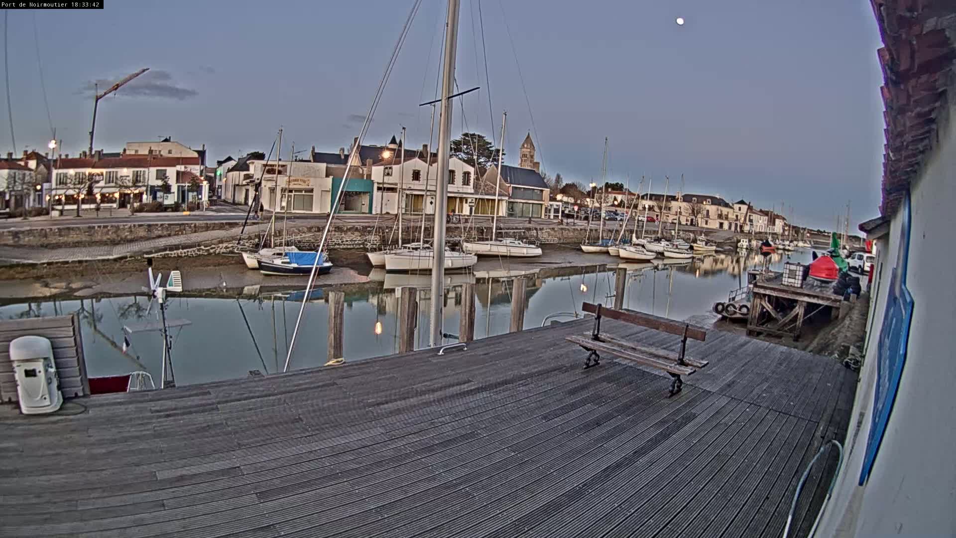 A calm harbor at dusk, under a clear sky and a visible moon, shows several sailboats moored along a stone quay, with a wooden dock in the foreground.