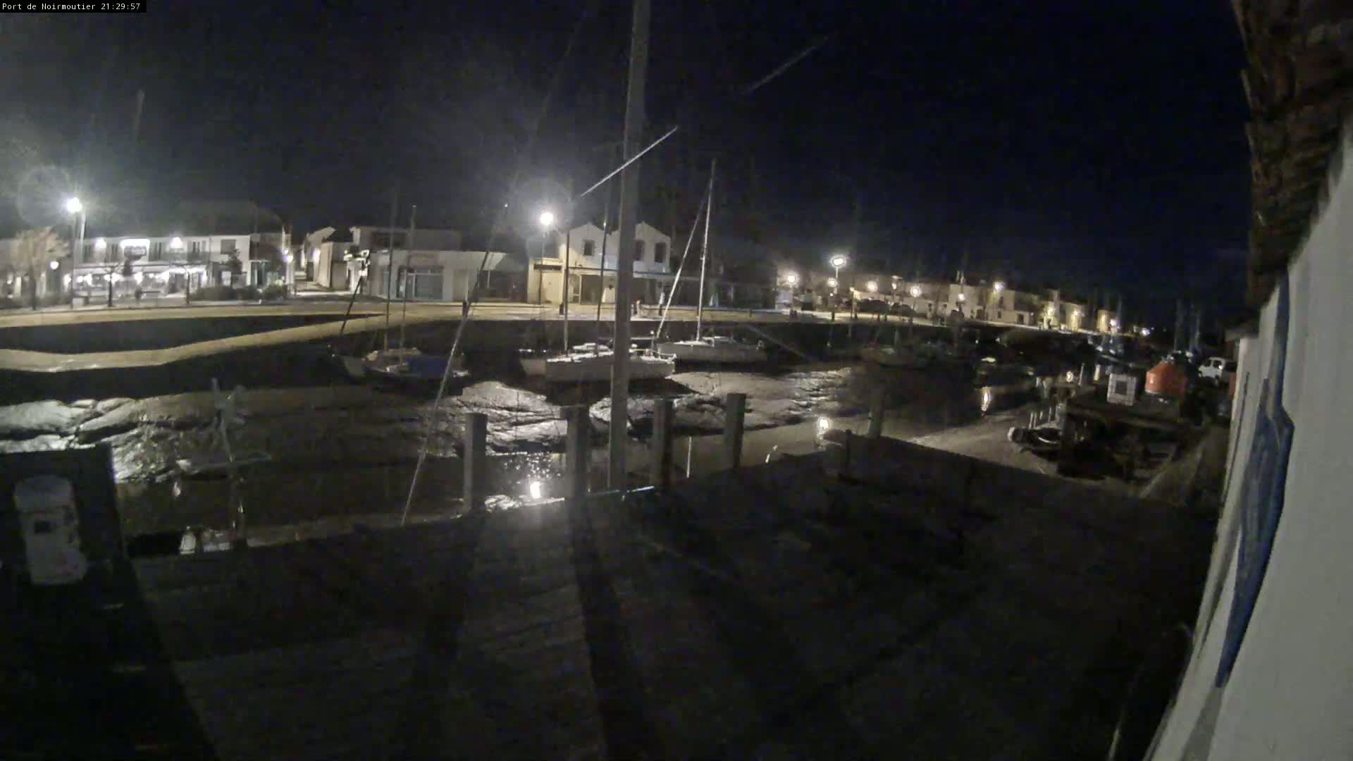 A nighttime view of a harbor with several boats docked at low tide, illuminated by streetlights along a waterfront building.