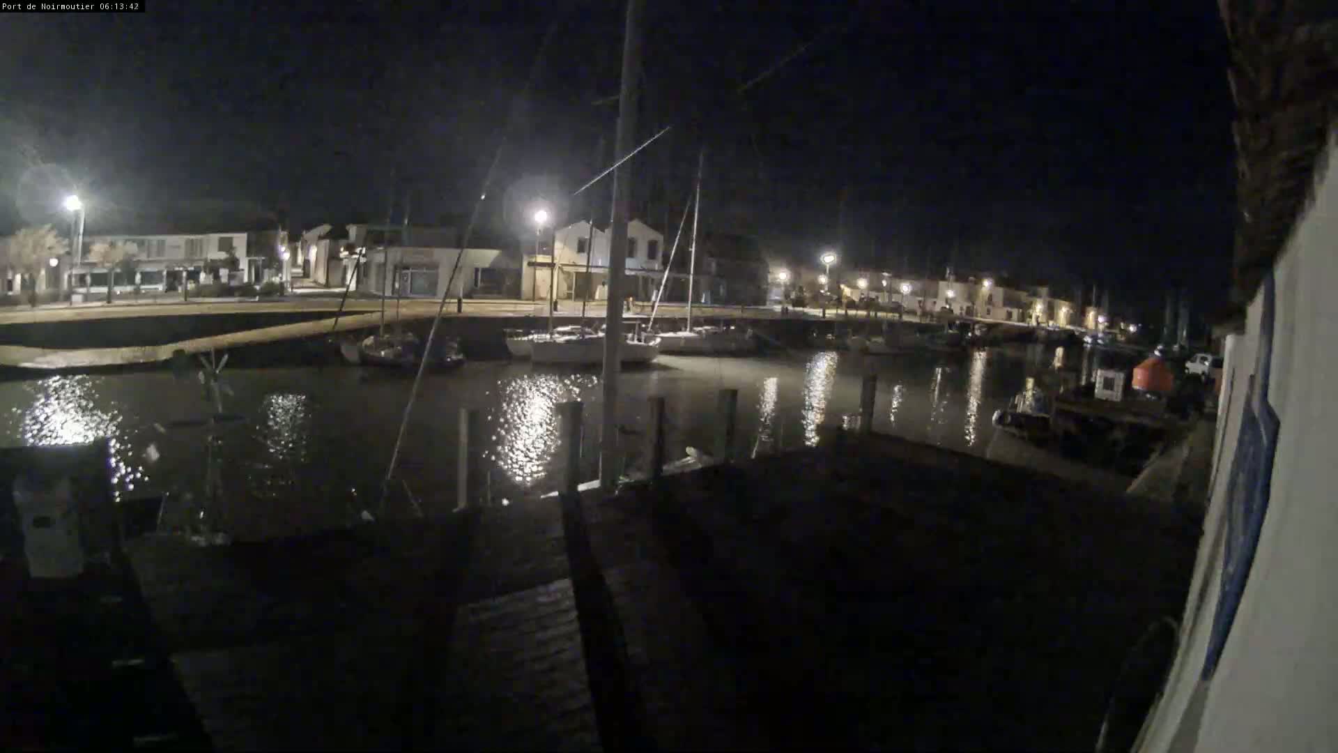At night, sailboats are moored in a calm harbor next to a waterfront town illuminated by streetlights.