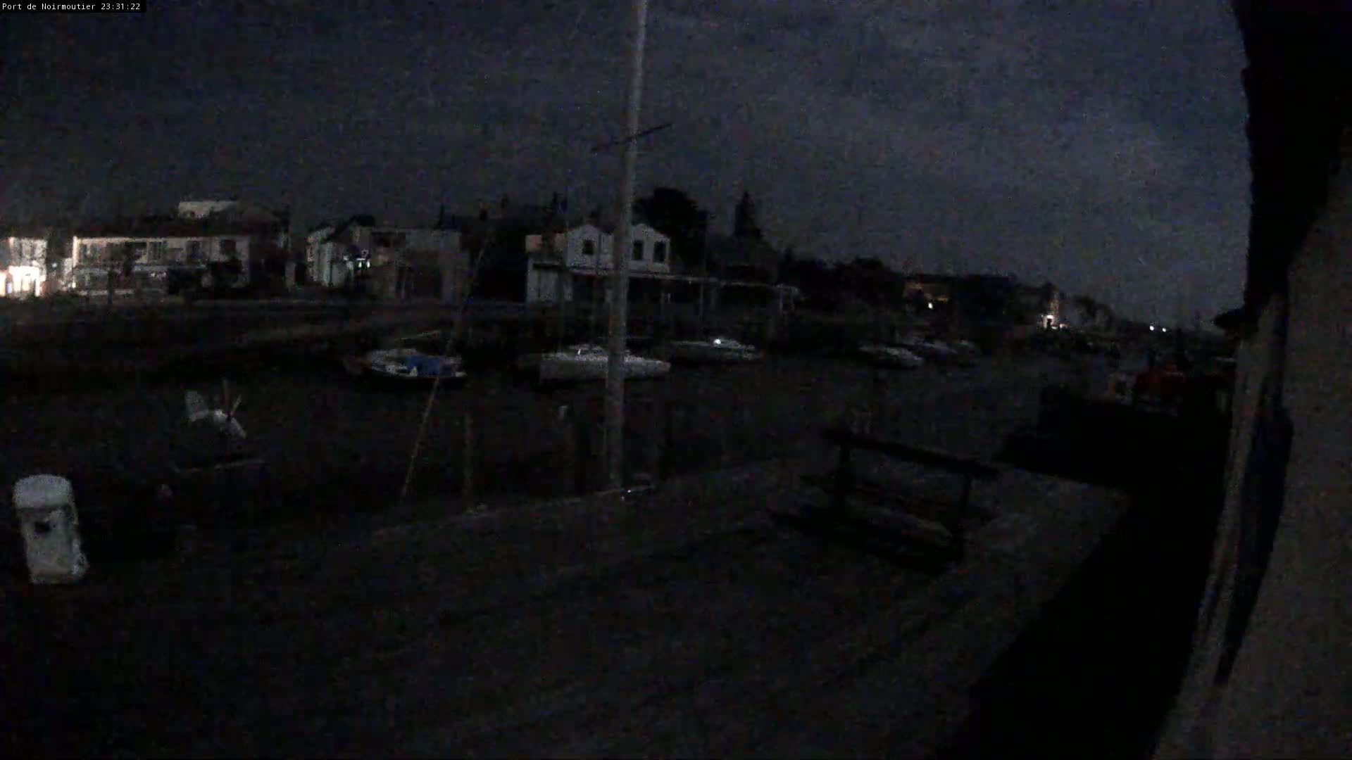 A dark nighttime view of a harbor with several boats docked near buildings under a cloudy sky.