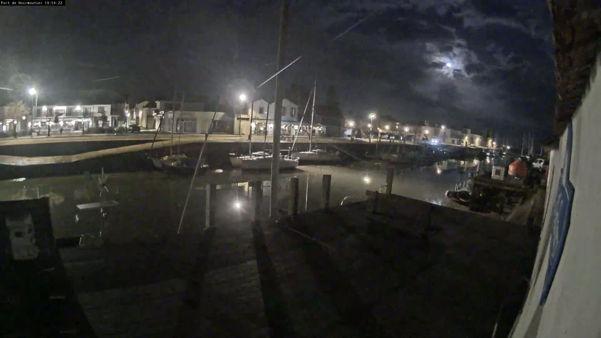 A nighttime view of a harbor with boats docked, buildings lining the waterfront under a partly cloudy moonlit sky.