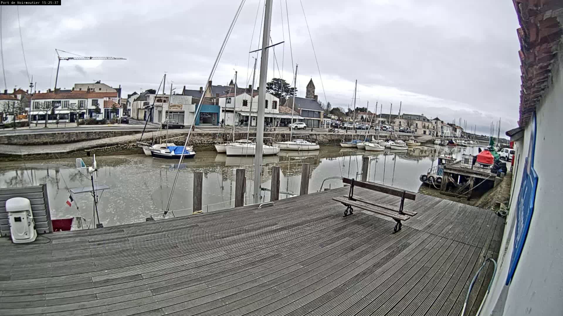 A wooden dock overlooks a calm harbor filled with sailboats moored alongside a waterfront town under an overcast sky.