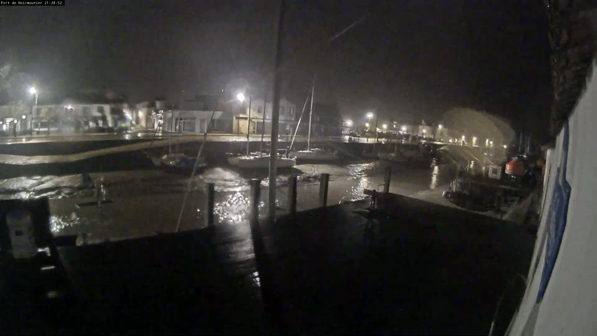 A nighttime view of a harbor with choppy water, several boats moored at the docks, and buildings lit by streetlights in the background during stormy weather.