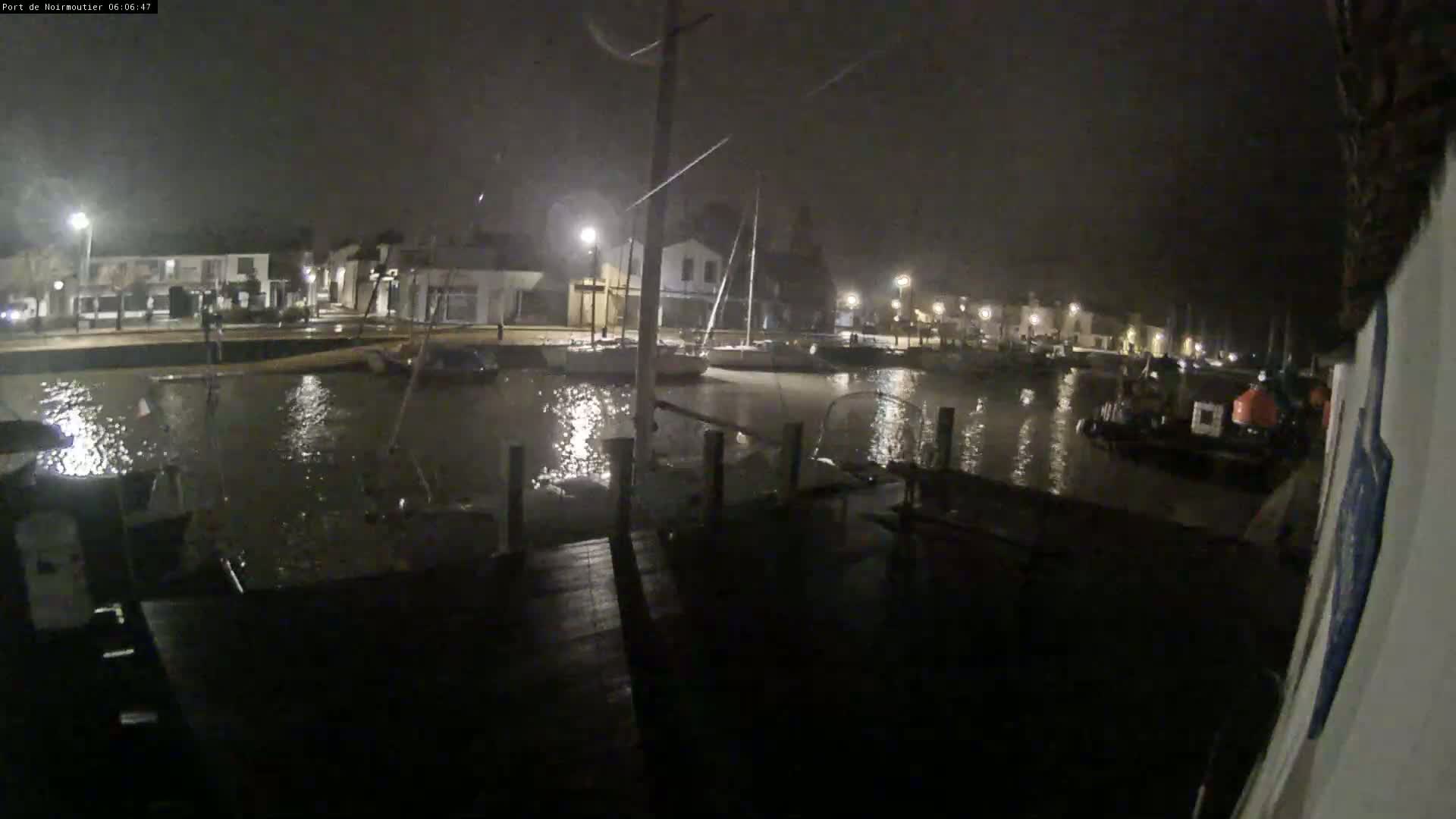 A nighttime view of a harbor with boats moored alongside a dock in rainy conditions.