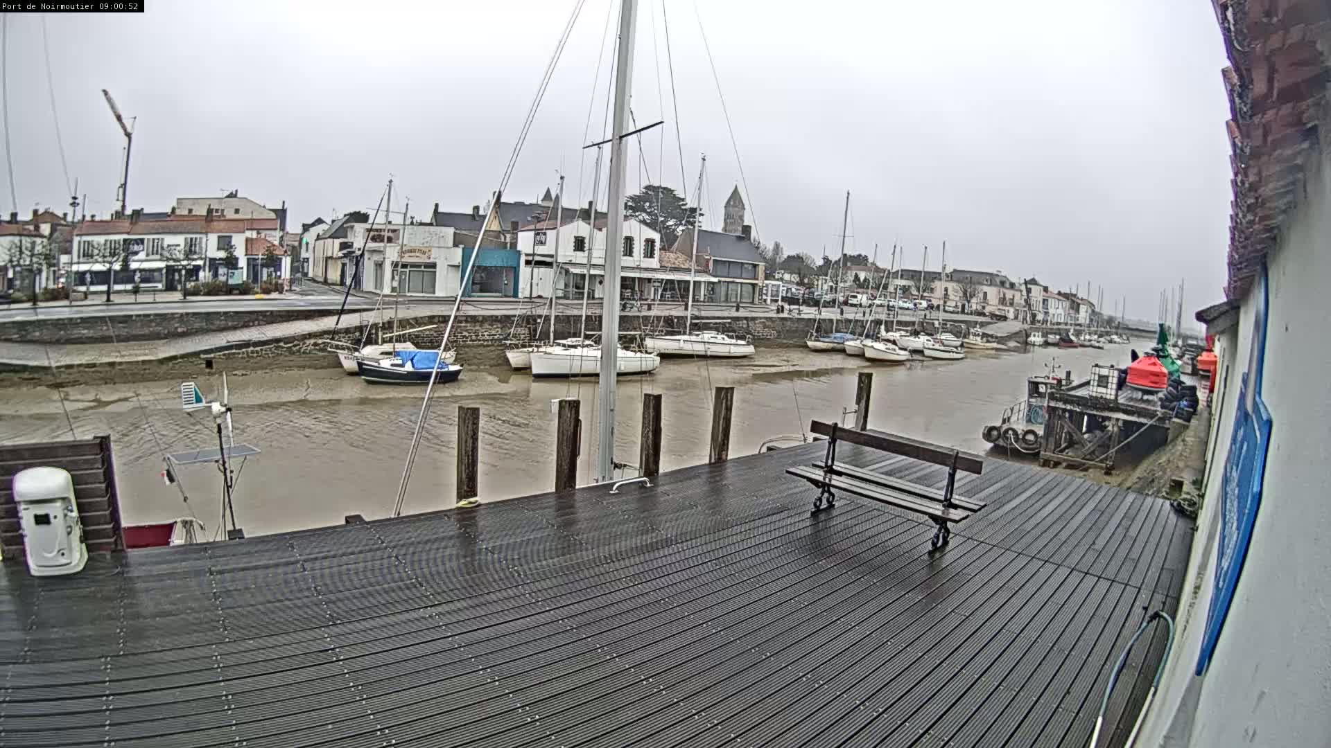 A wet wooden dock overlooks a harbor with several sailboats at low tide, under an overcast sky.