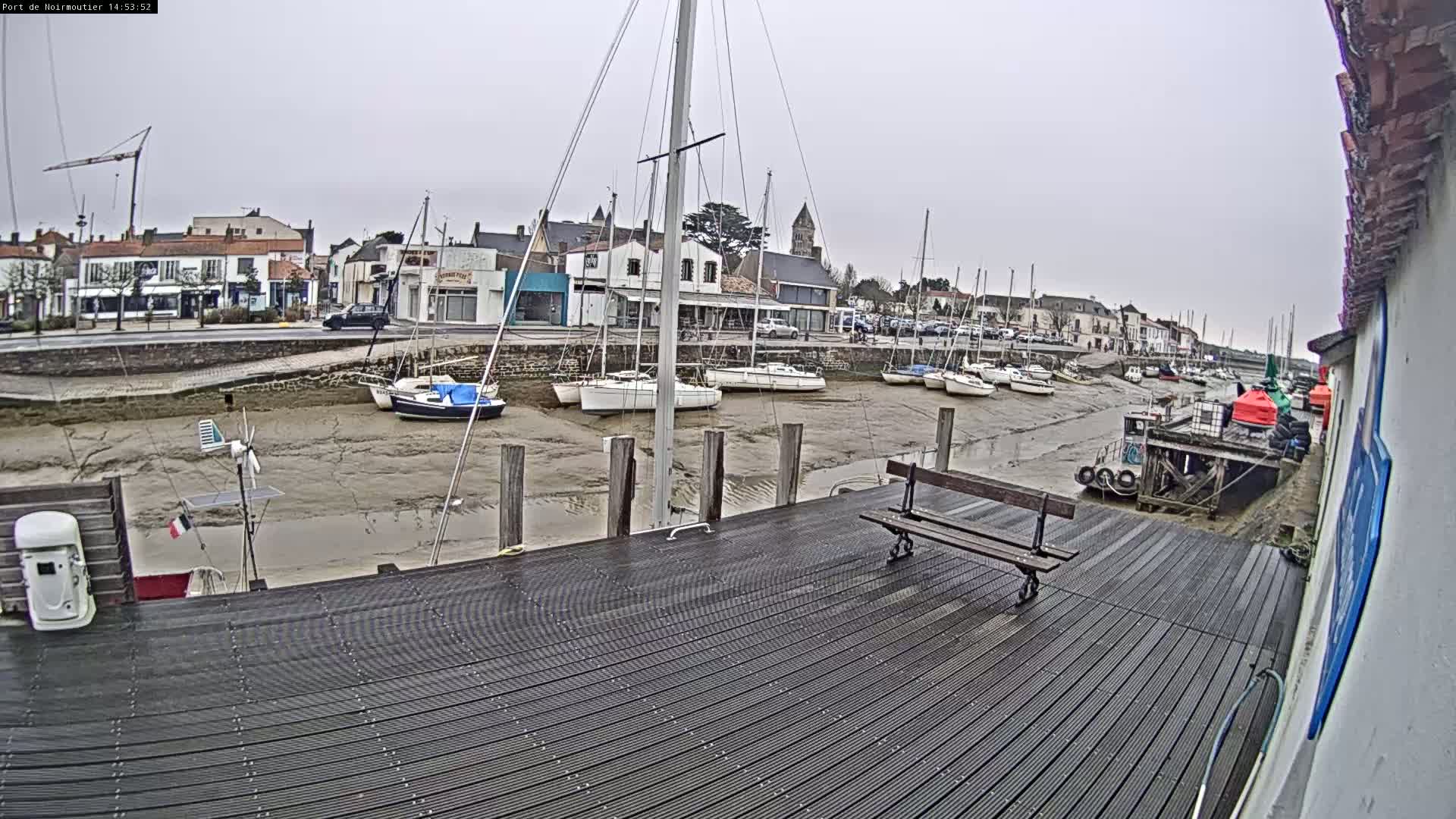 A harbor with several sailboats docked on a muddy, low-tide waterfront, viewed from a dark wooden dock on an overcast day.