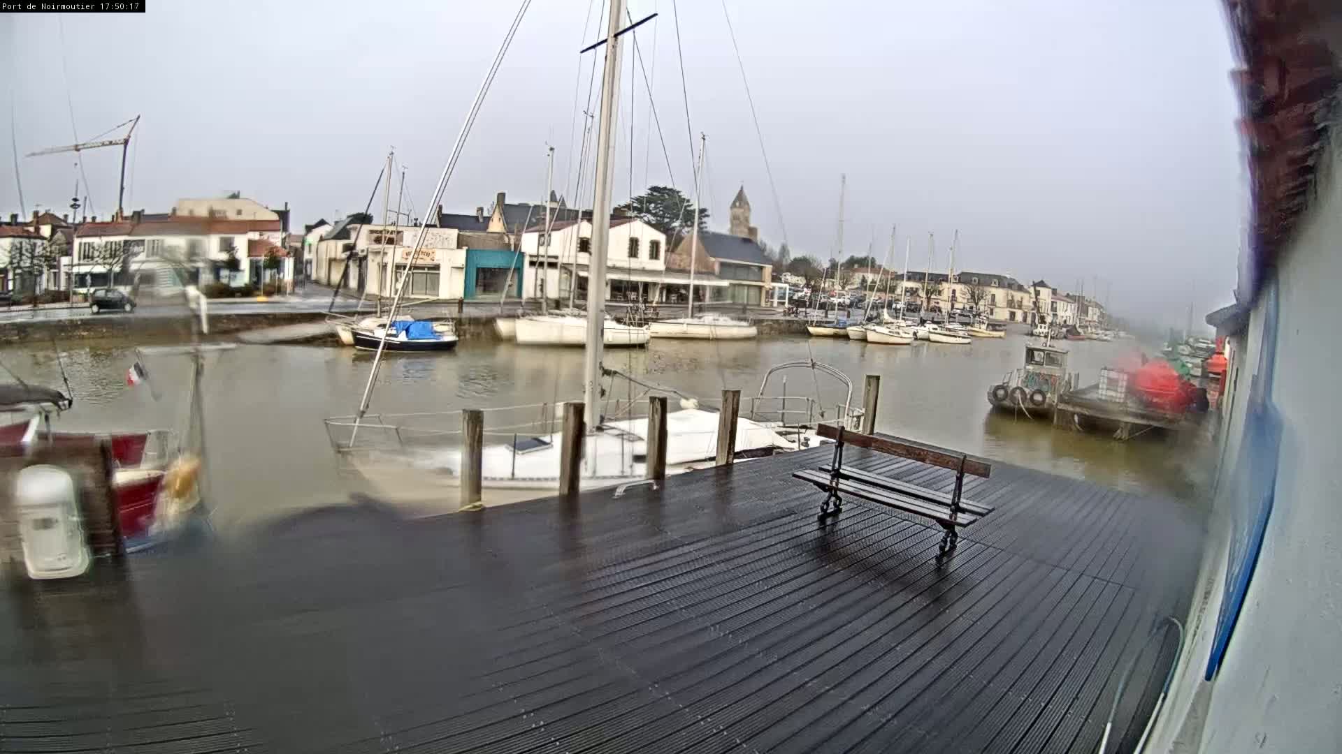 A wet dockside with several sailboats moored, viewed from a dark wooden platform under an overcast sky.