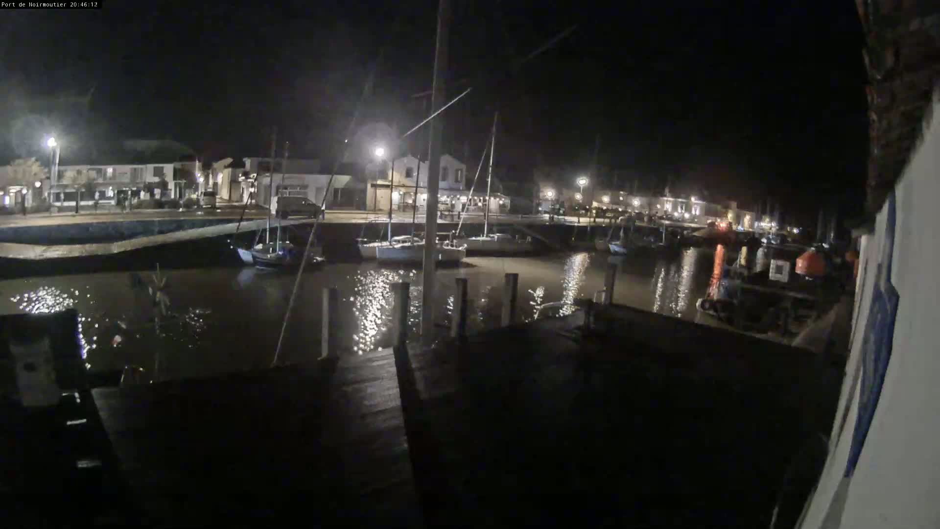 A nighttime view of a harbor with several sailboats moored, illuminated by streetlights reflecting on the calm water.