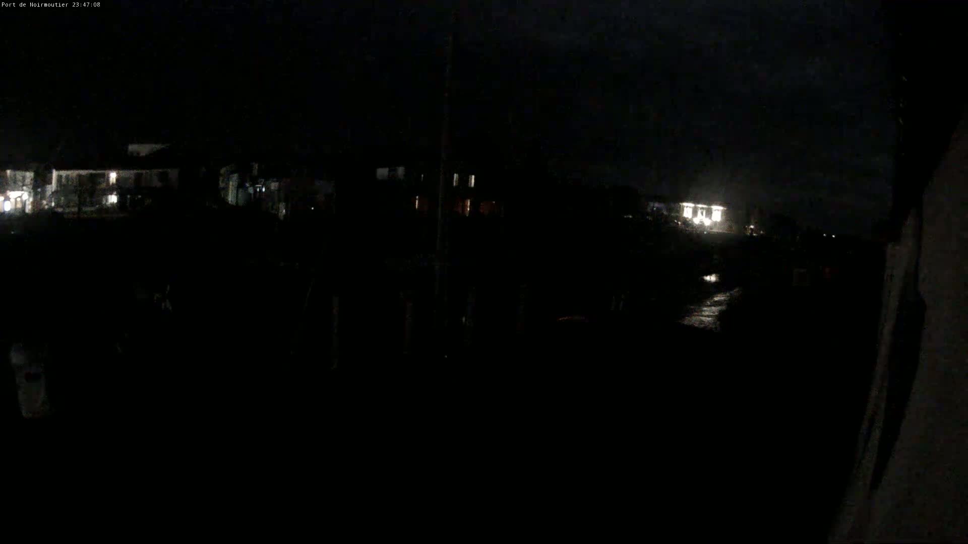 A dark nighttime scene shows dimly lit buildings and a wet, reflective surface in the foreground under a cloudy sky.